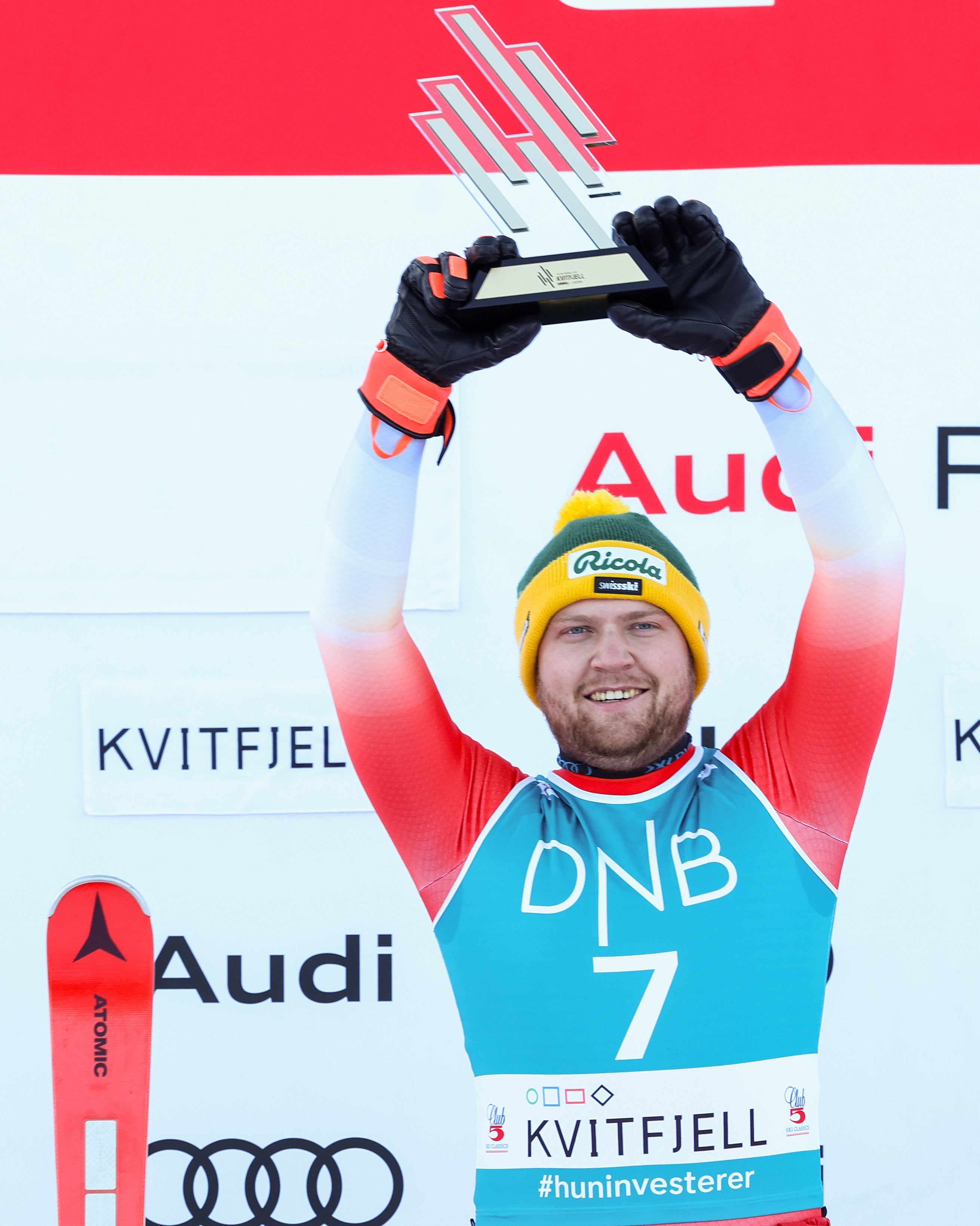 Niels Hintermann a red a ski jersey with the number 7, holding a silver trophy above his head on a podium. He is wearing a yellow and green beanie, black gloves, and red ski pants.