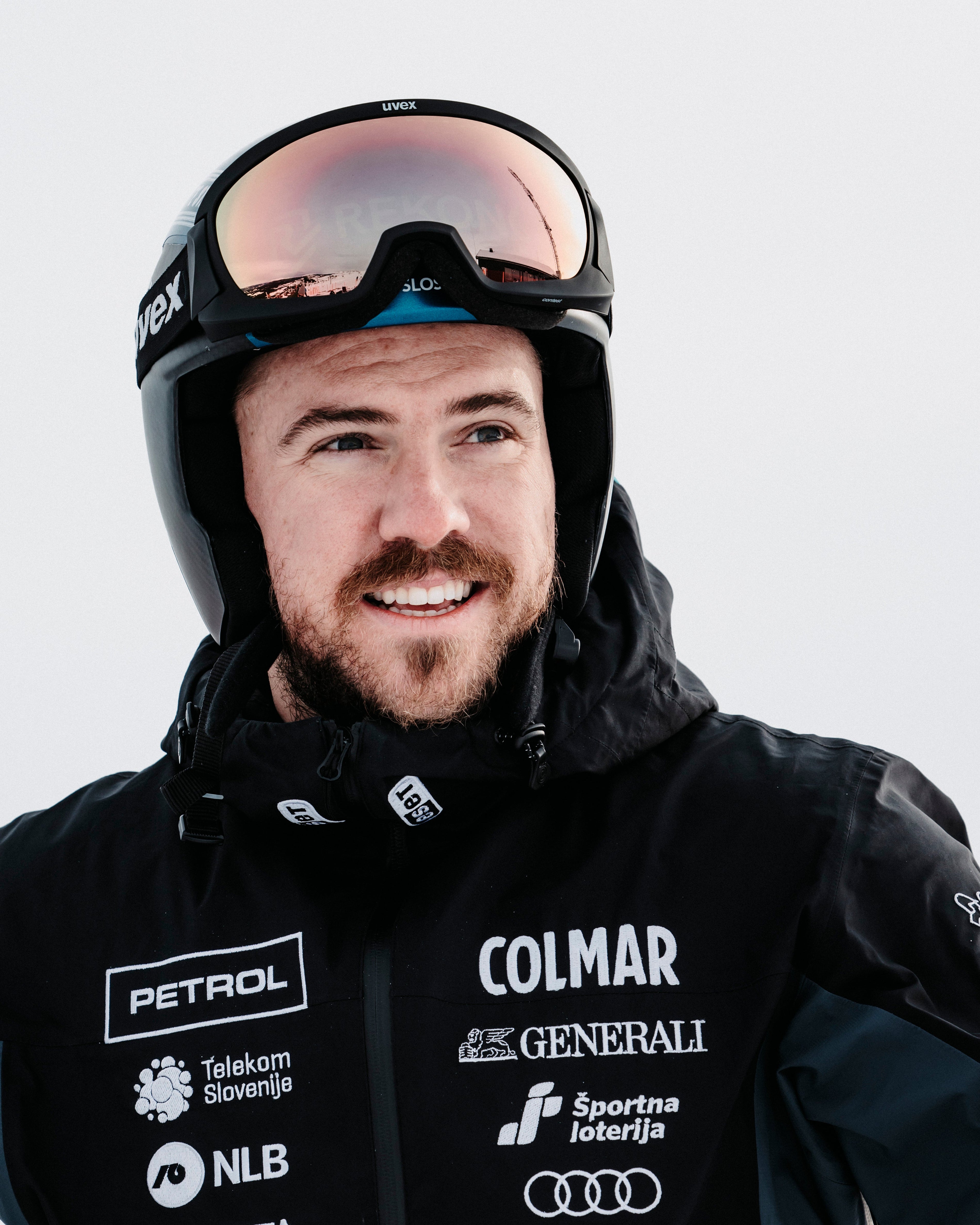 A close-up portrait of a smiling Miha Hrobat wearing a black ski helmet with mirrored goggles and a black ski jacket covered in sponsor logos, set against a bright, overexposed snowy background.