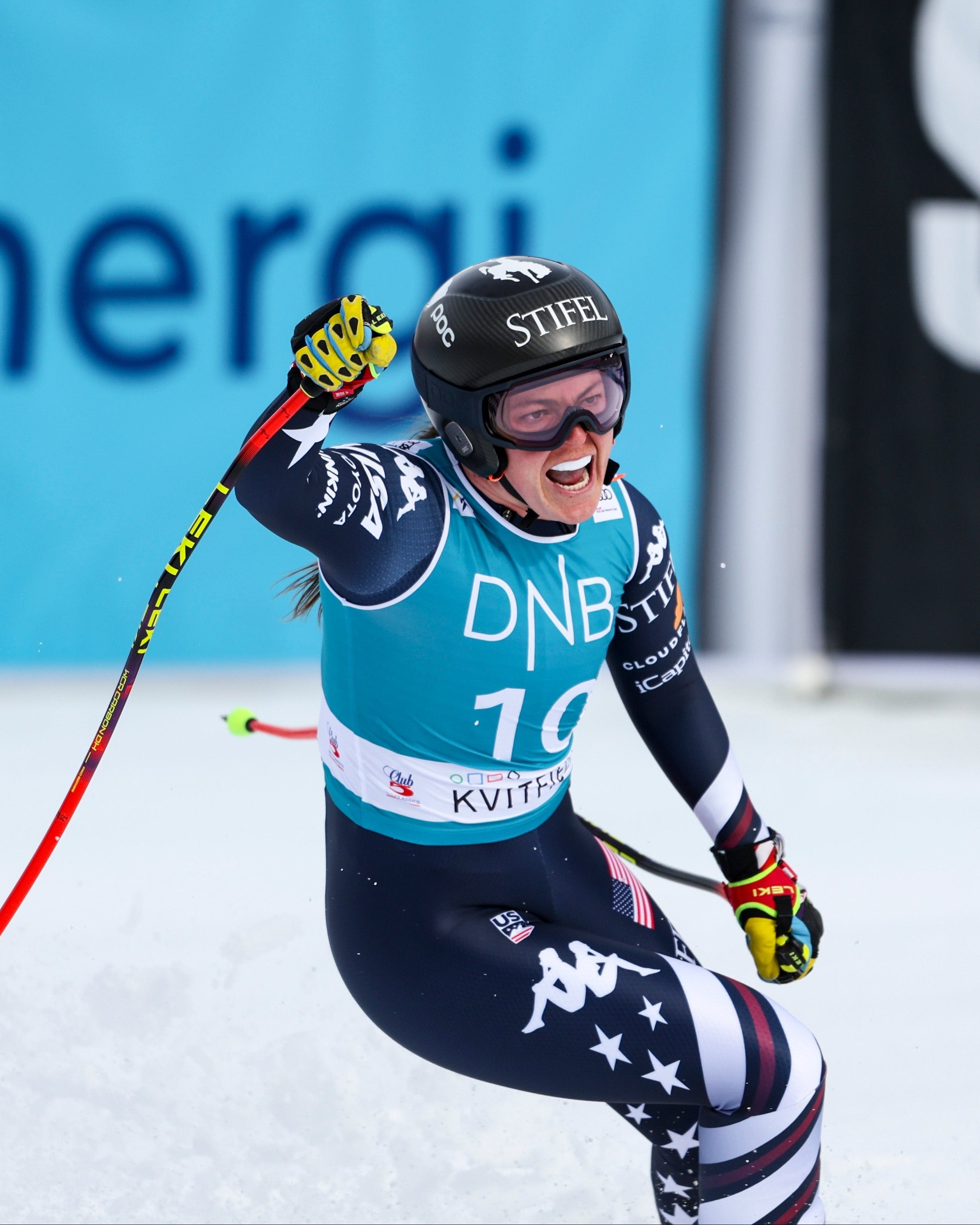 Breezy Johnson in a dark blue, white, and red USA team suit celebrates after a race on a snowy track, wearing a black helmet and goggles.