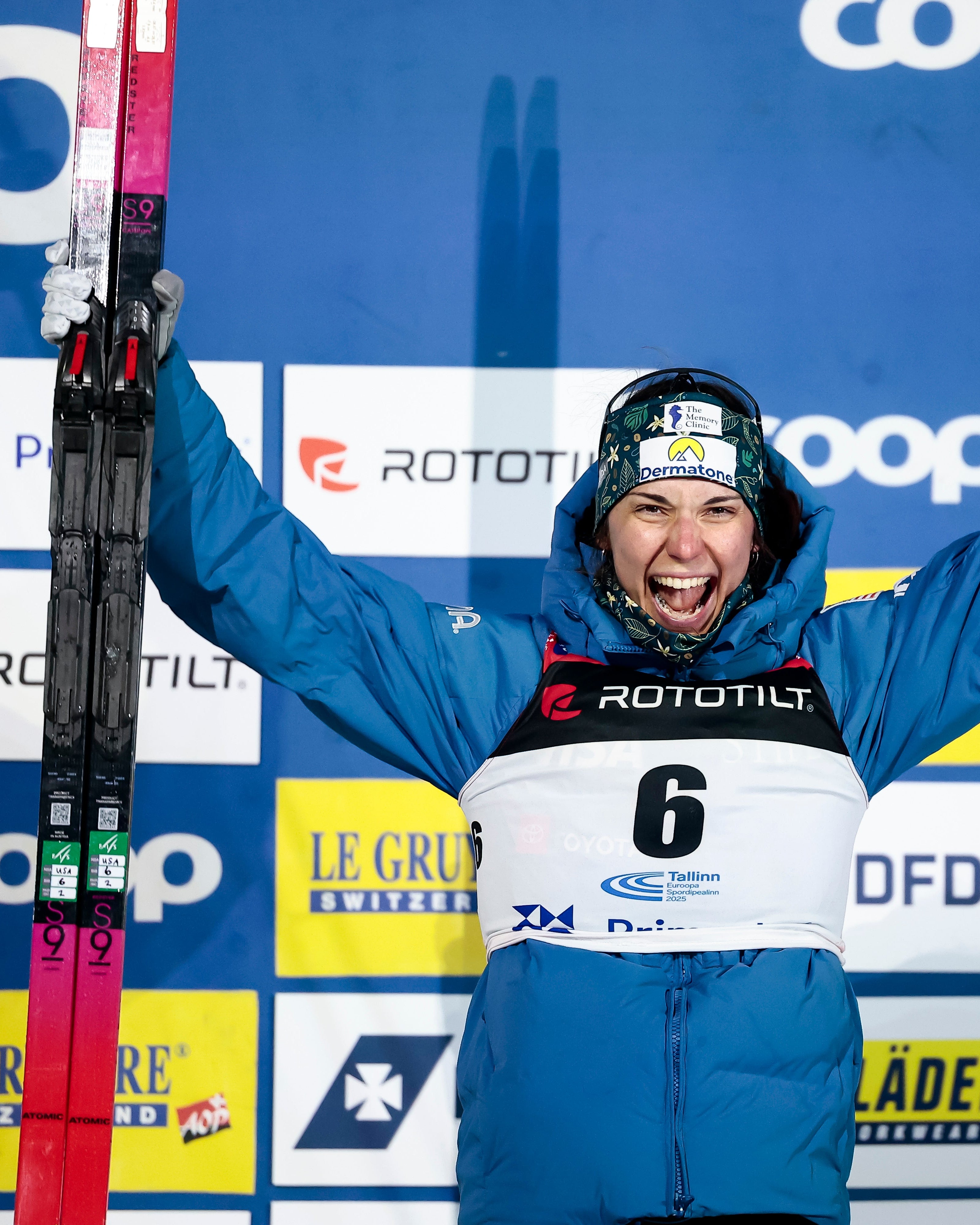 Julia Kern celebrates on the podium with arms raised, wearing a blue ski suit with a white bib numbered 6 and holding ski poles and skis in front of a blue and yellow sponsor backdrop.
