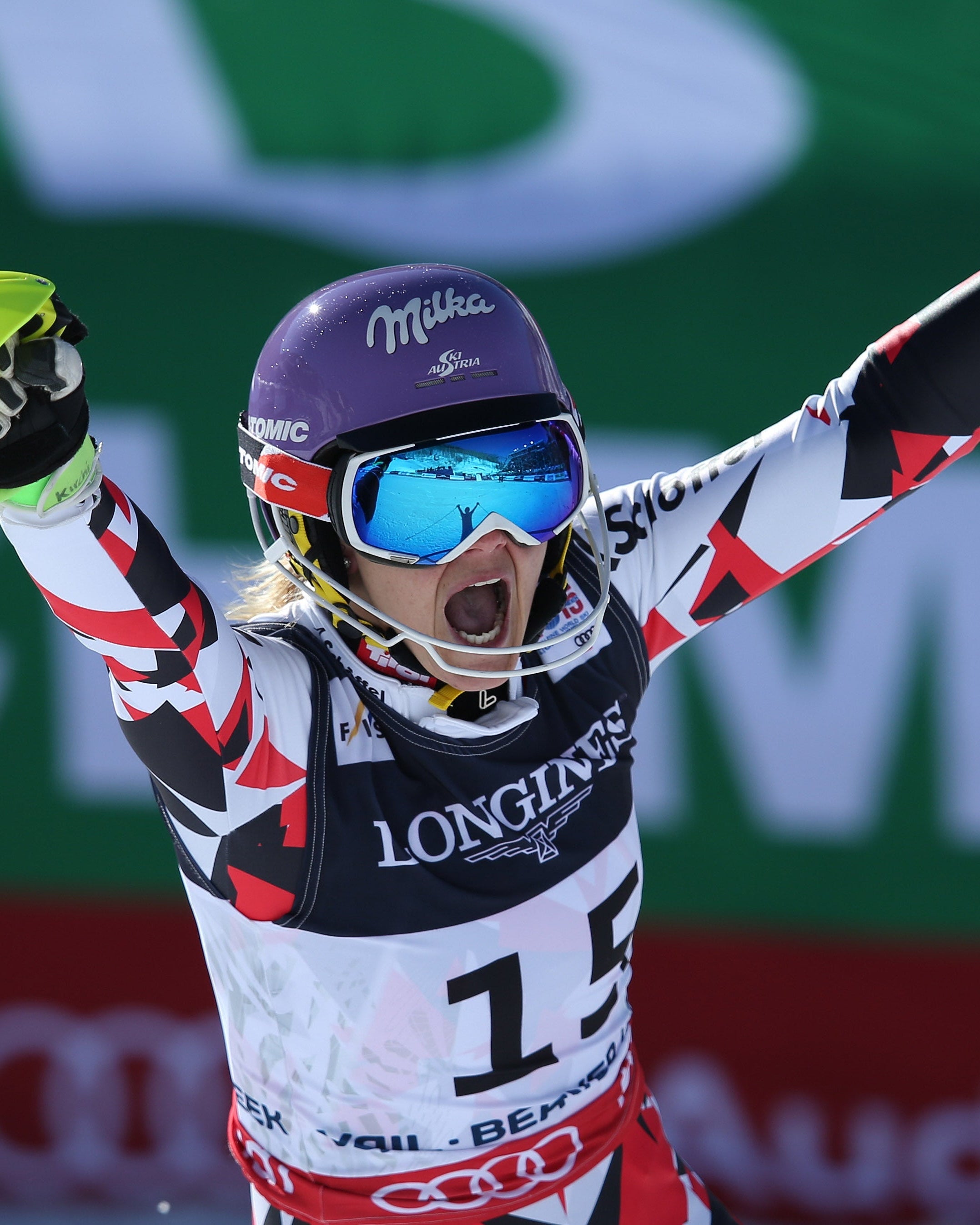 Michaela Kirchgasser celebrates on the slopes with arms raised, wearing a white, red, and black racing suit, a purple helmet, blue goggles, with a green and white event backdrop behind her.