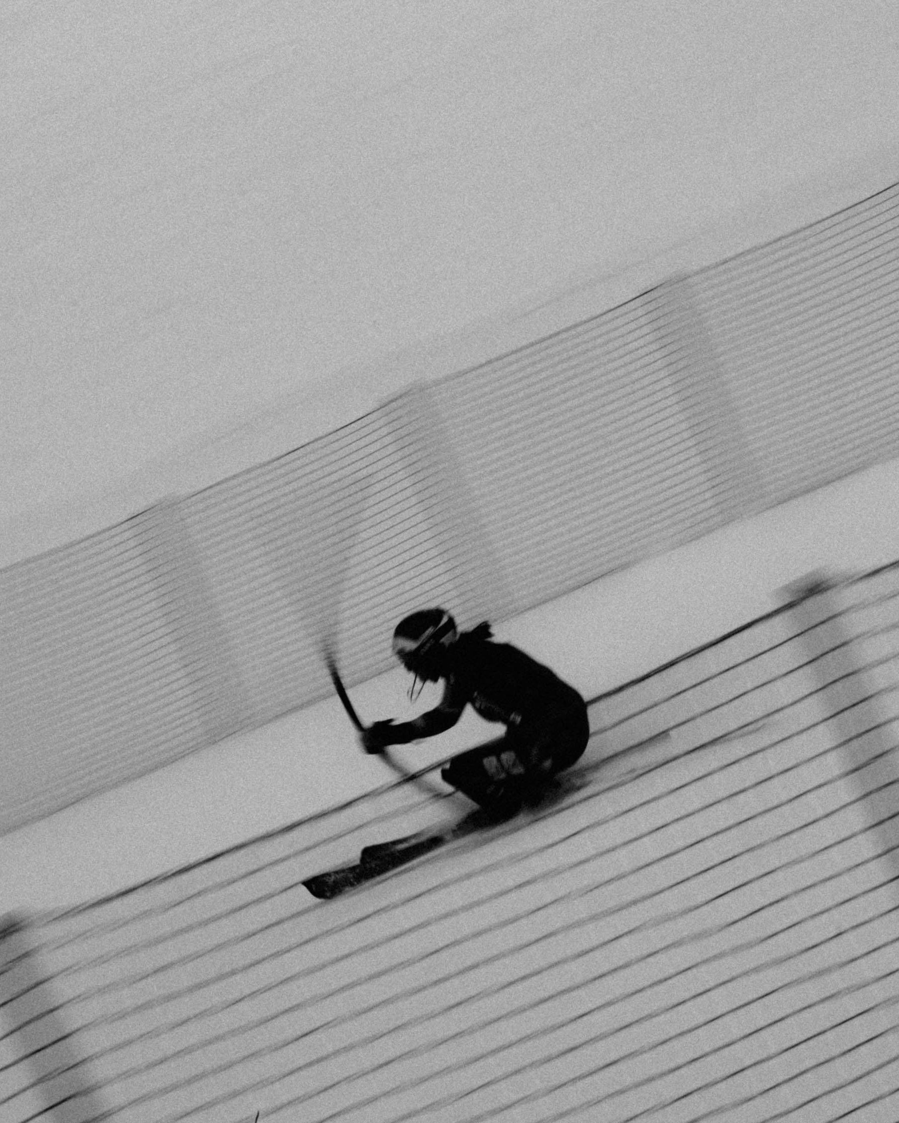 A black-and-white action shot of Zrinka Ljutic in dark gear sliding down a snowy slalom slope beside a safety net, captured with a dynamic diagonal composition.