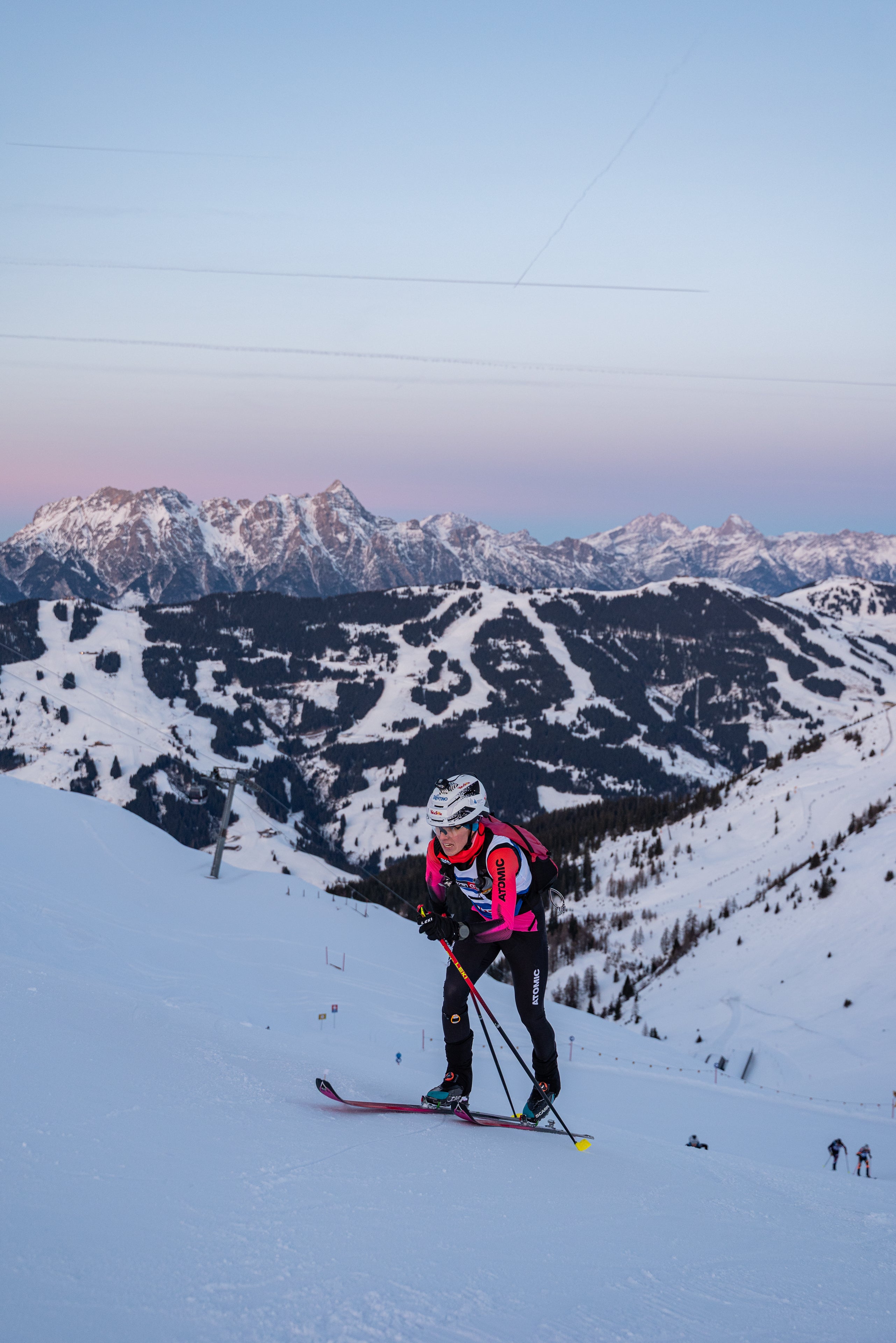 Davide Magnini in a bright pink and black jacket stands on a snowy mountain slope with a dramatic backdrop of snow-covered peaks under a pale blue and pink sky.
