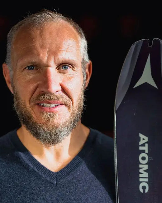 Hermann Maier wearing a dark blue V-neck sweater, is shown in a close-up portrait next to a black Atomic ski with white branding.