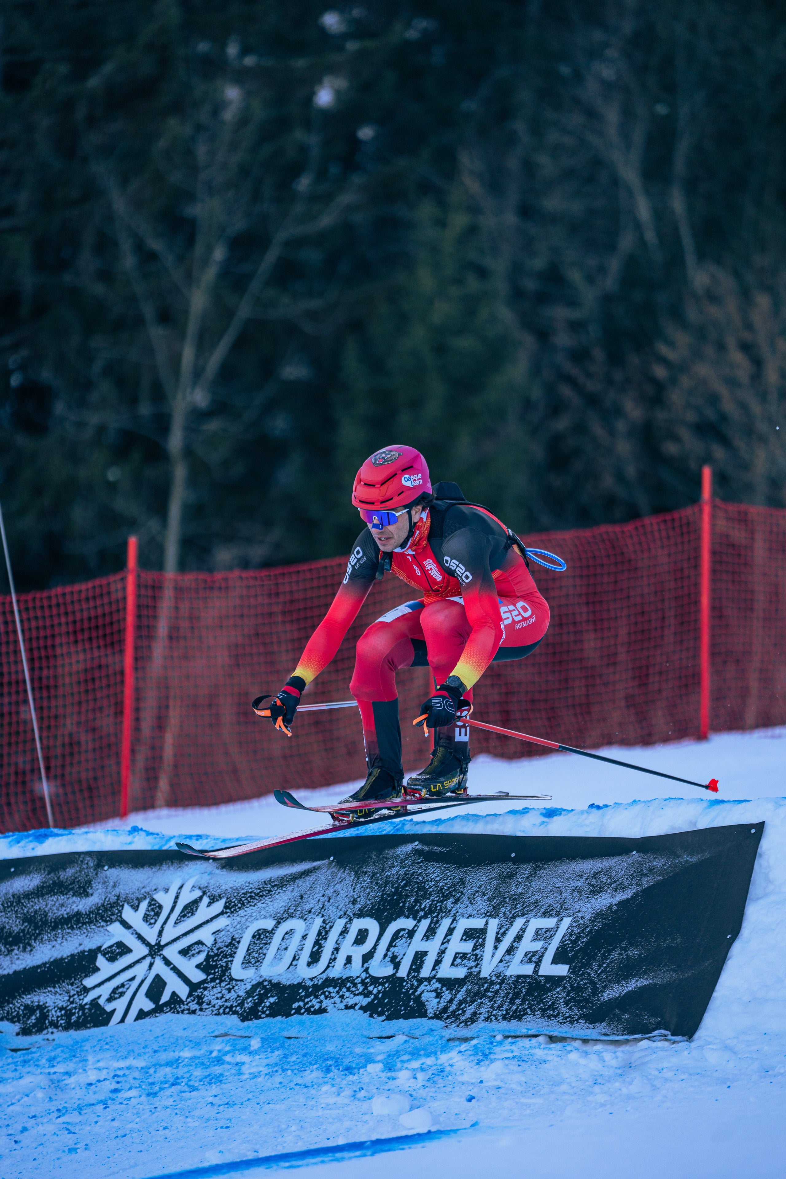 Íñigo Martínez de Albornoz in a red and black racing suit jumps over a black snowboard ramp branded COURGHEVEL, with snow, trees, and a red safety net in the background.