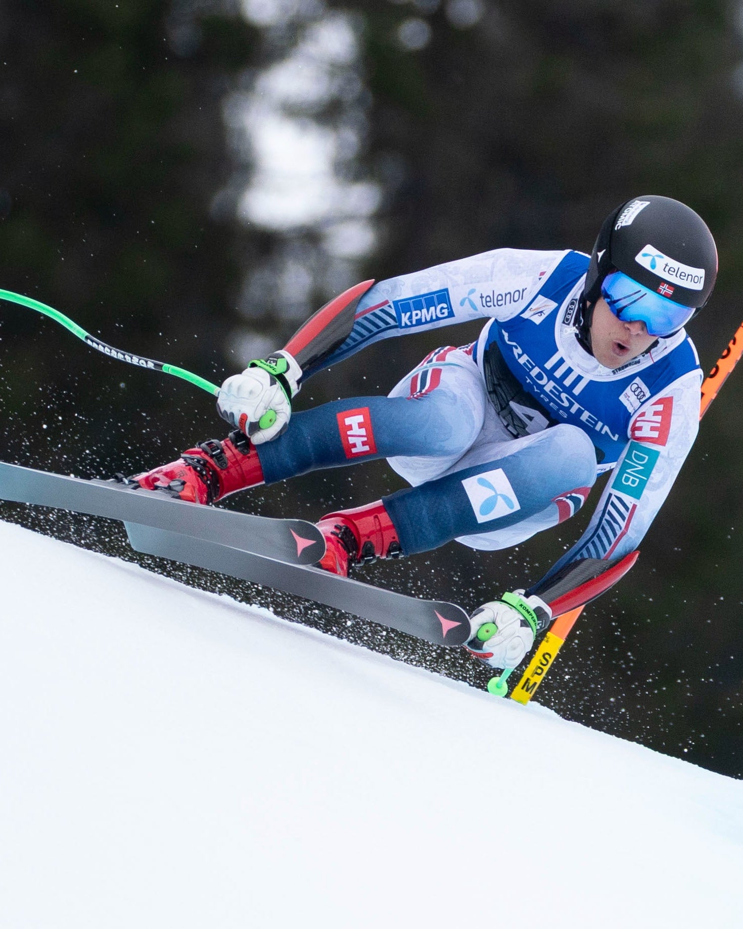 A skier in a blue, white, and red racing suit leans sharply into a turn on a snowy slope, with an orange slalom gate and dark pine trees in the background.