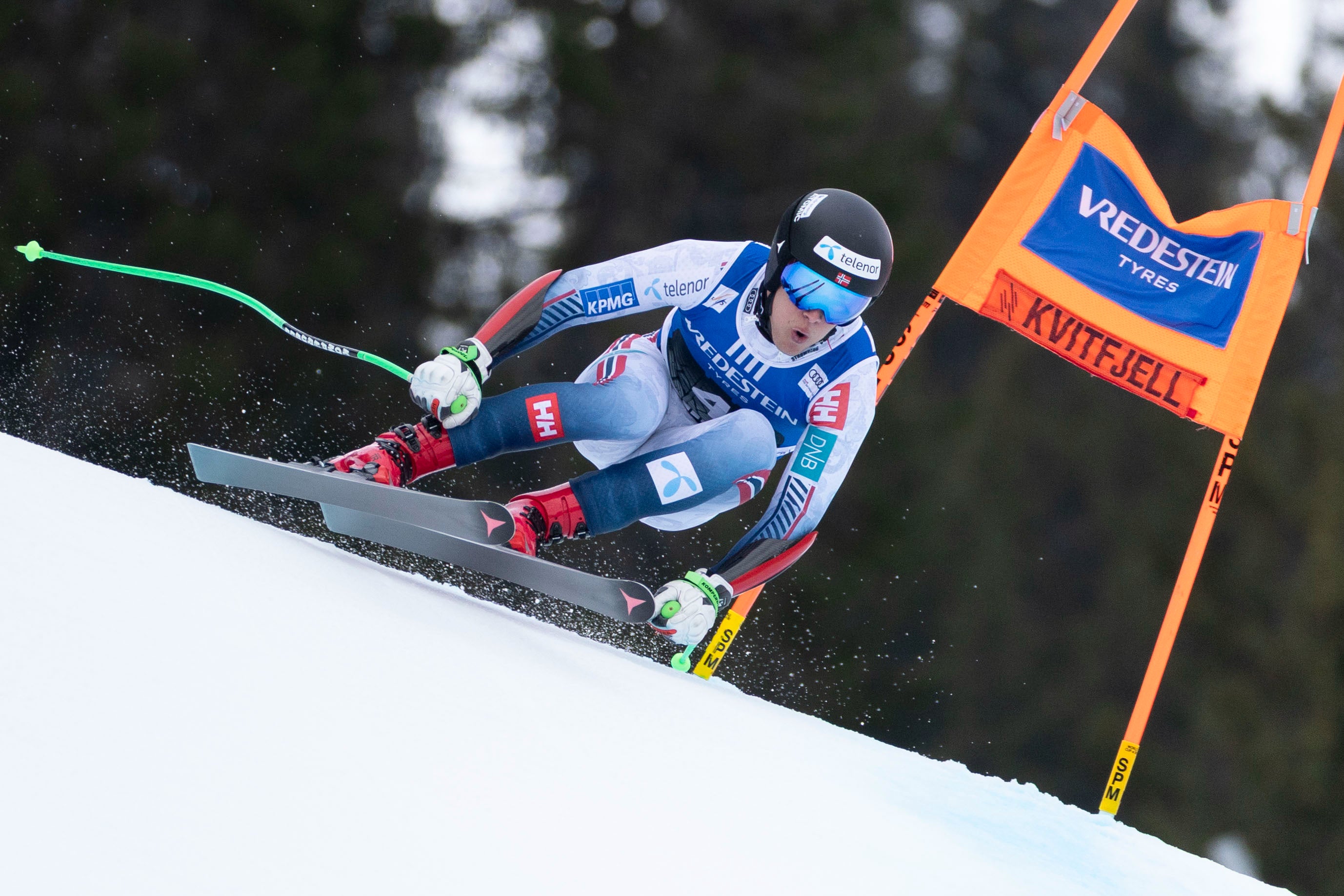 A skier in a blue, white, and red racing suit leans sharply into a turn on a snowy slope, with an orange slalom gate and dark pine trees in the background.