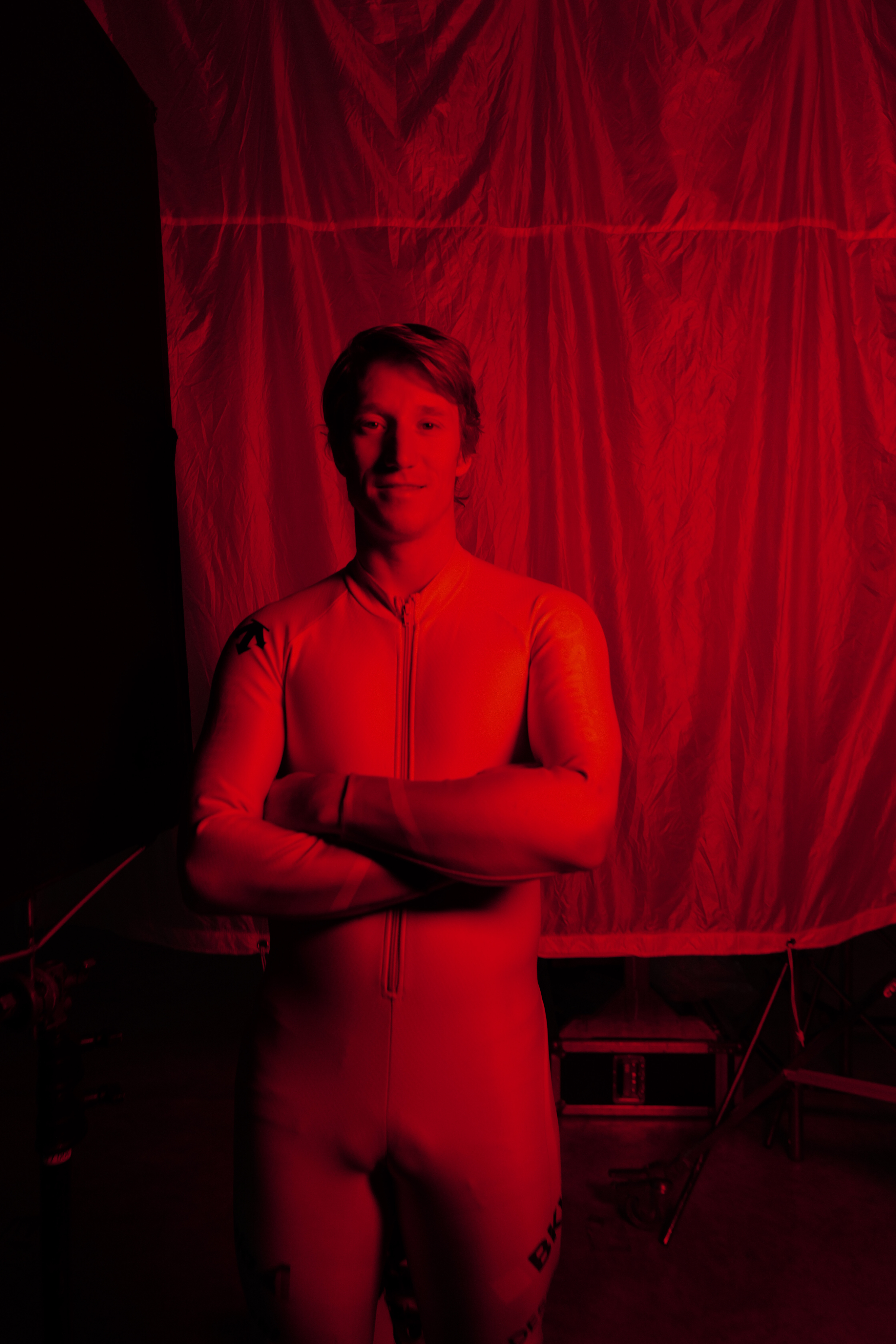 Tanguy Nef wearing a full-body white or off-white one-piece suit or bodysuit stands with arms crossed in front of a red-lit backdrop. The scene is heavily tinted red, and the background appears to be a studio setup with draped fabric and equipment.