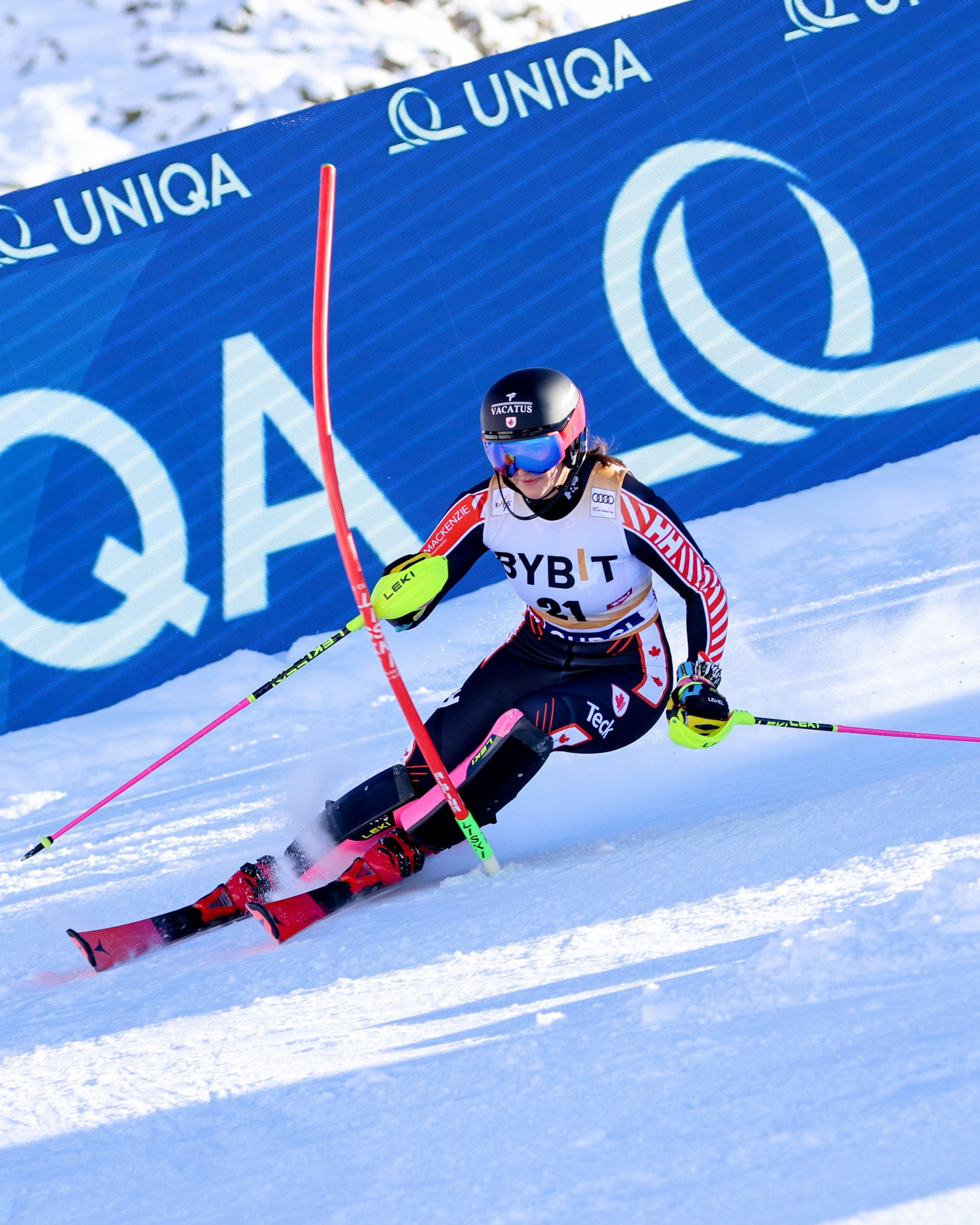 Ali Nullmeyer in a black, white, and red racing suit leans sharply into a turn on a snowy slope, holding ski poles in front of a large blue UNIQA advertising banner.