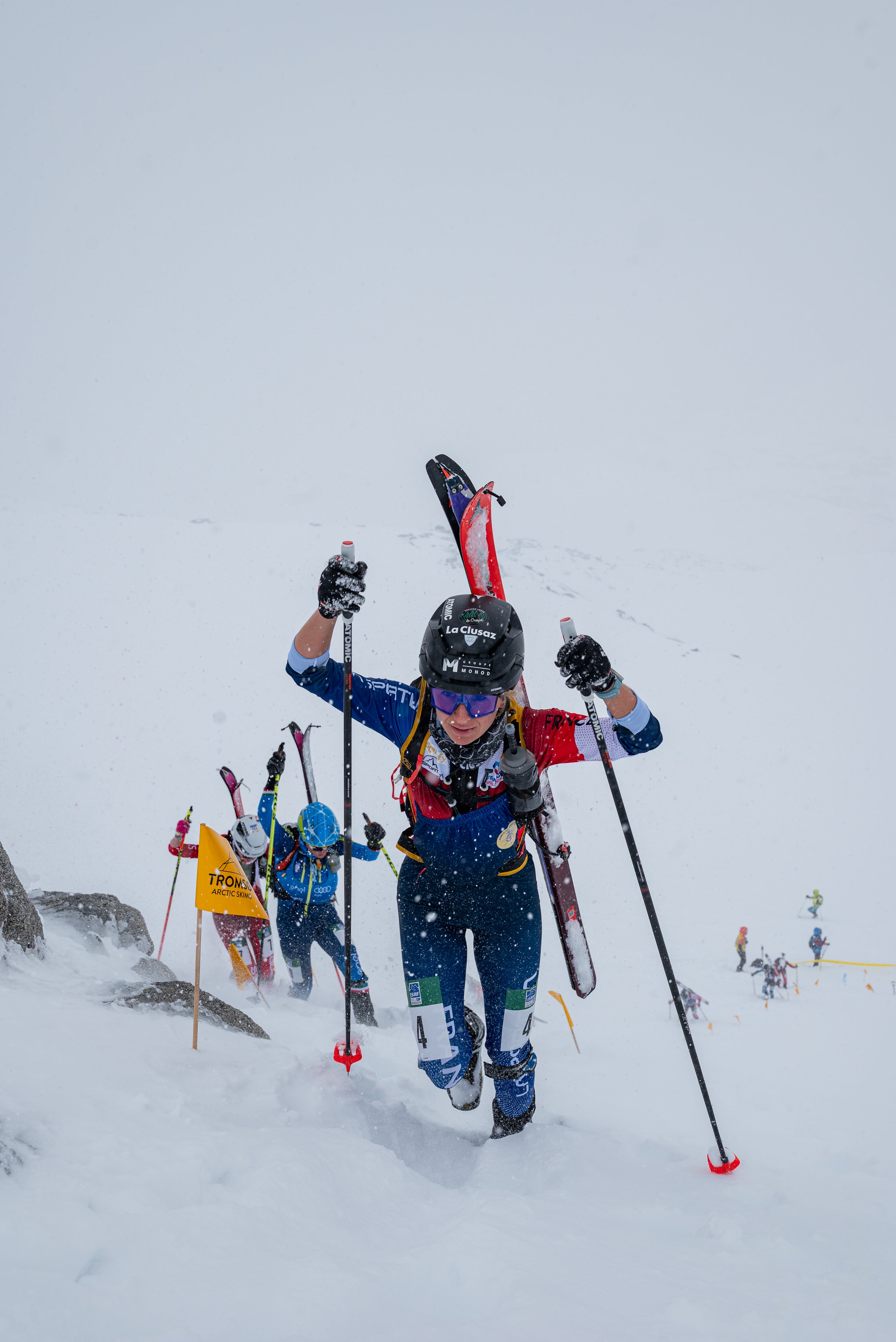 Celia Passey in a blue and red ski suit carries skis and poles while climbing through deep snow on a mountain, with other skiers visible in the background under an overcast sky.