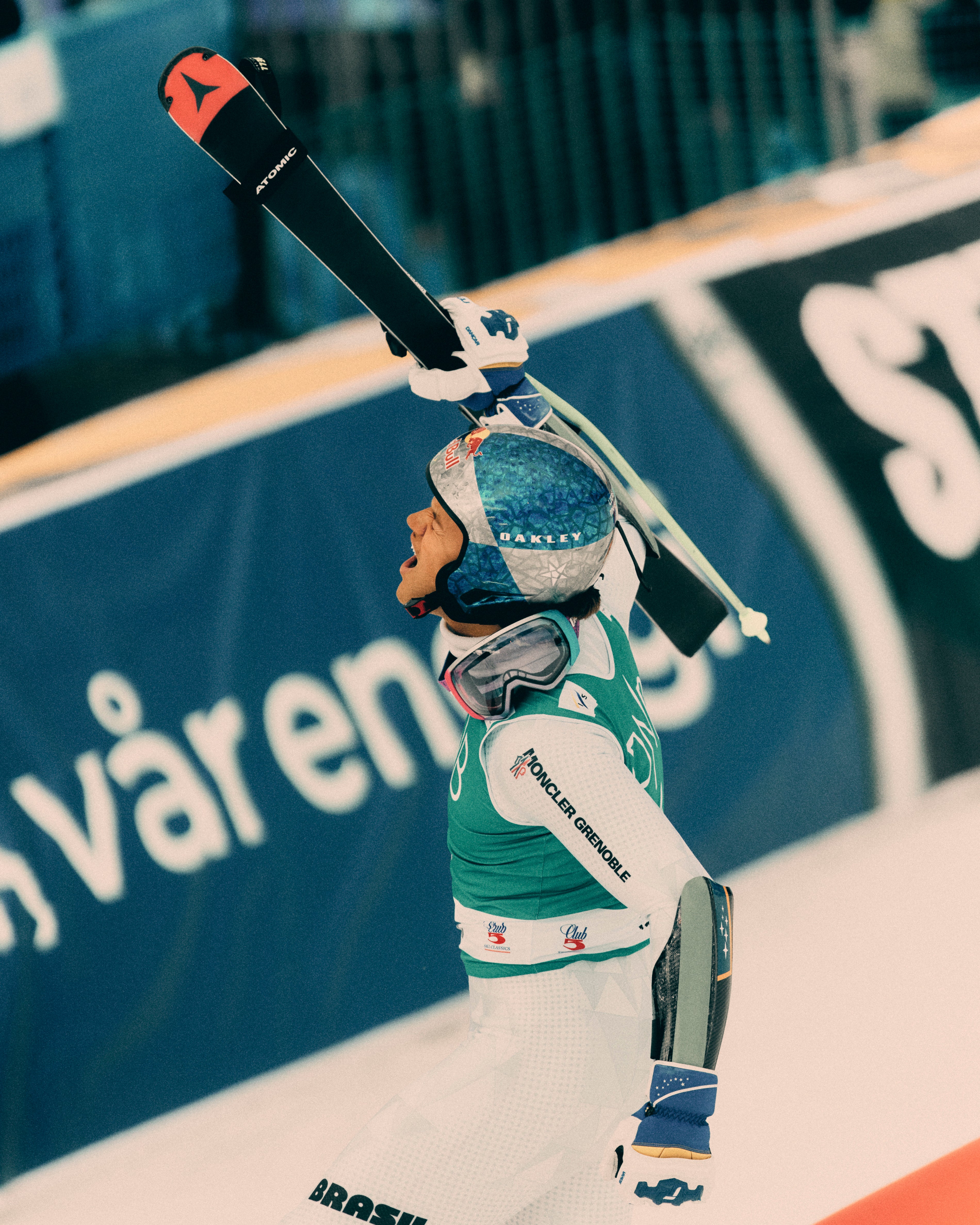 Lucas Pinheiro Braathen in a white ski suit with a blue helmet raises a ski above their head in a celebratory pose, with a blue advertising banner in the background.