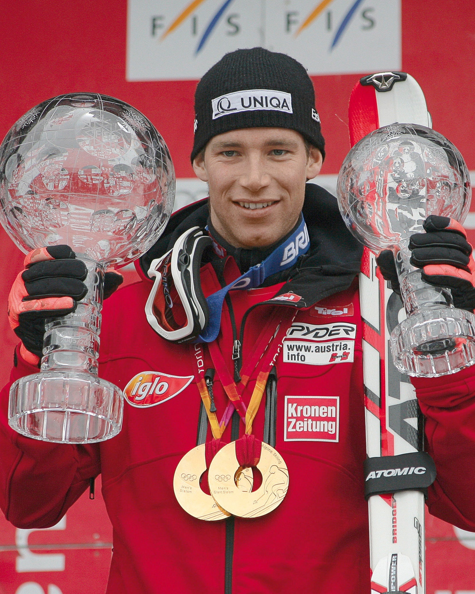 Benjamin Raich stands on a podium holding two large clear trophy cups, wearing a red ski jacket, black gloves, a black beanie and gold medals. 