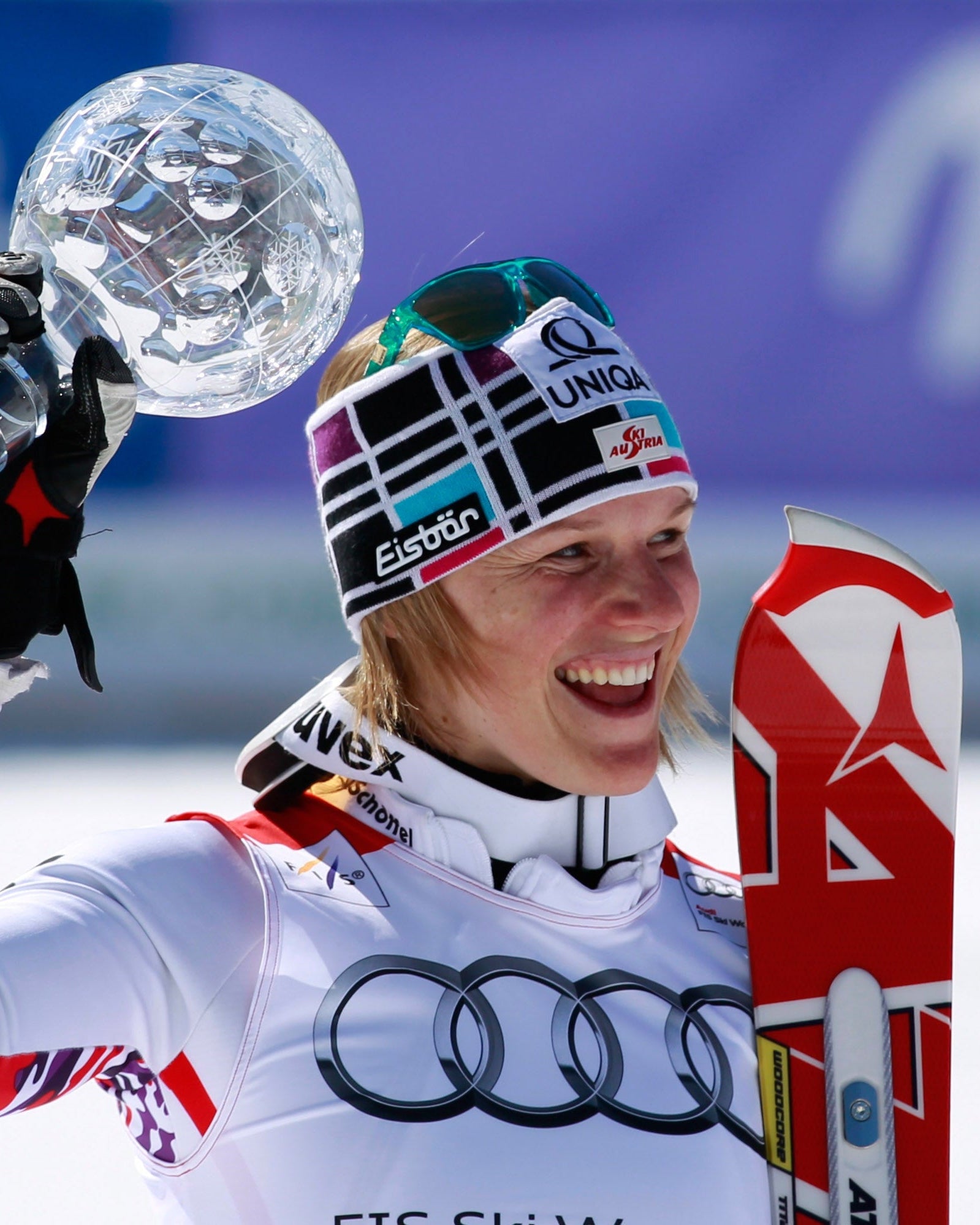A smiley Marlies Raich in a white racing suit and a colorful beanie holds up a clear crystal trophy above her head on a snowy outdoor track.