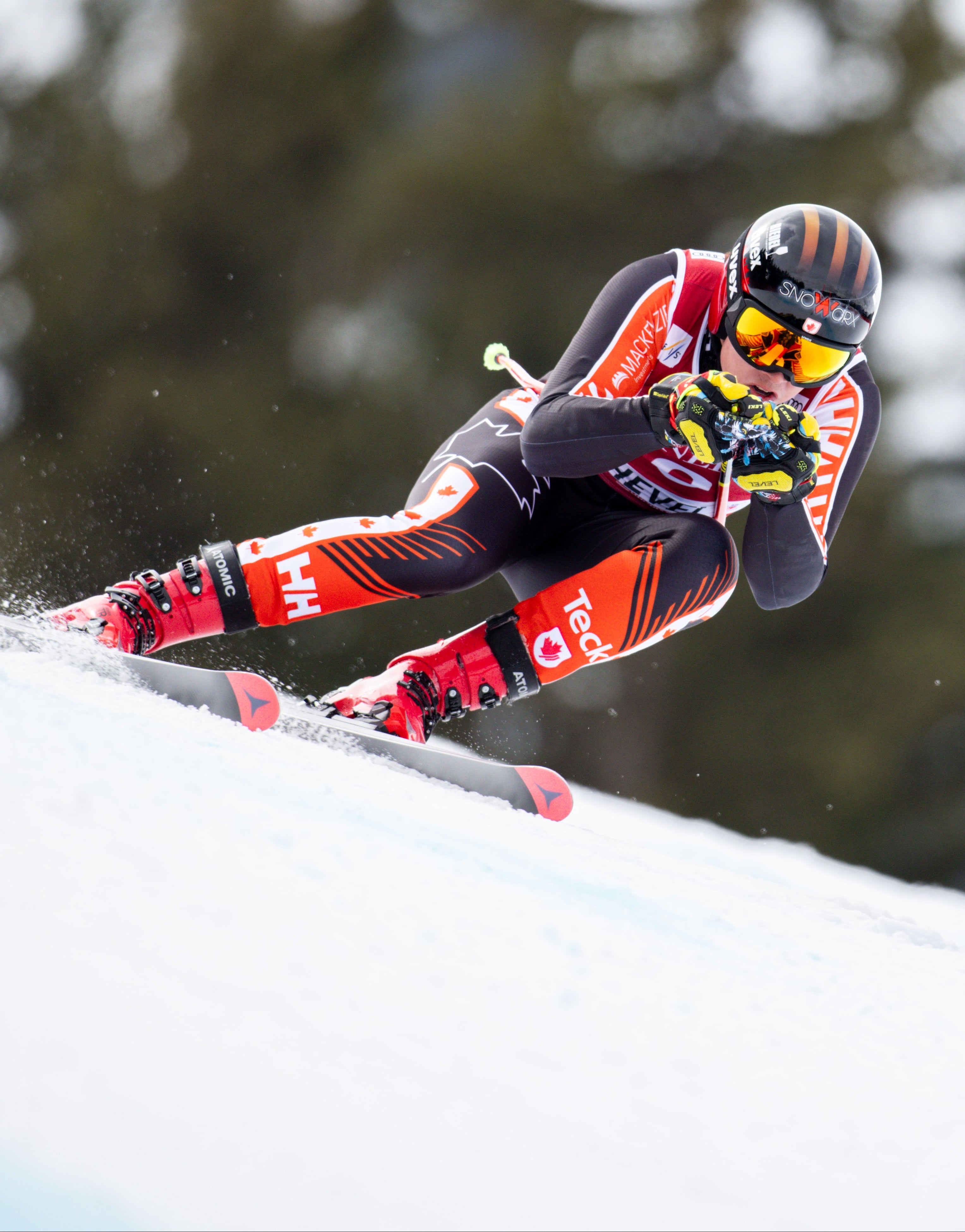 Jeffrey Read in a red, black, and orange racing suit leans sharply into a snowy turn on a mountain slope.