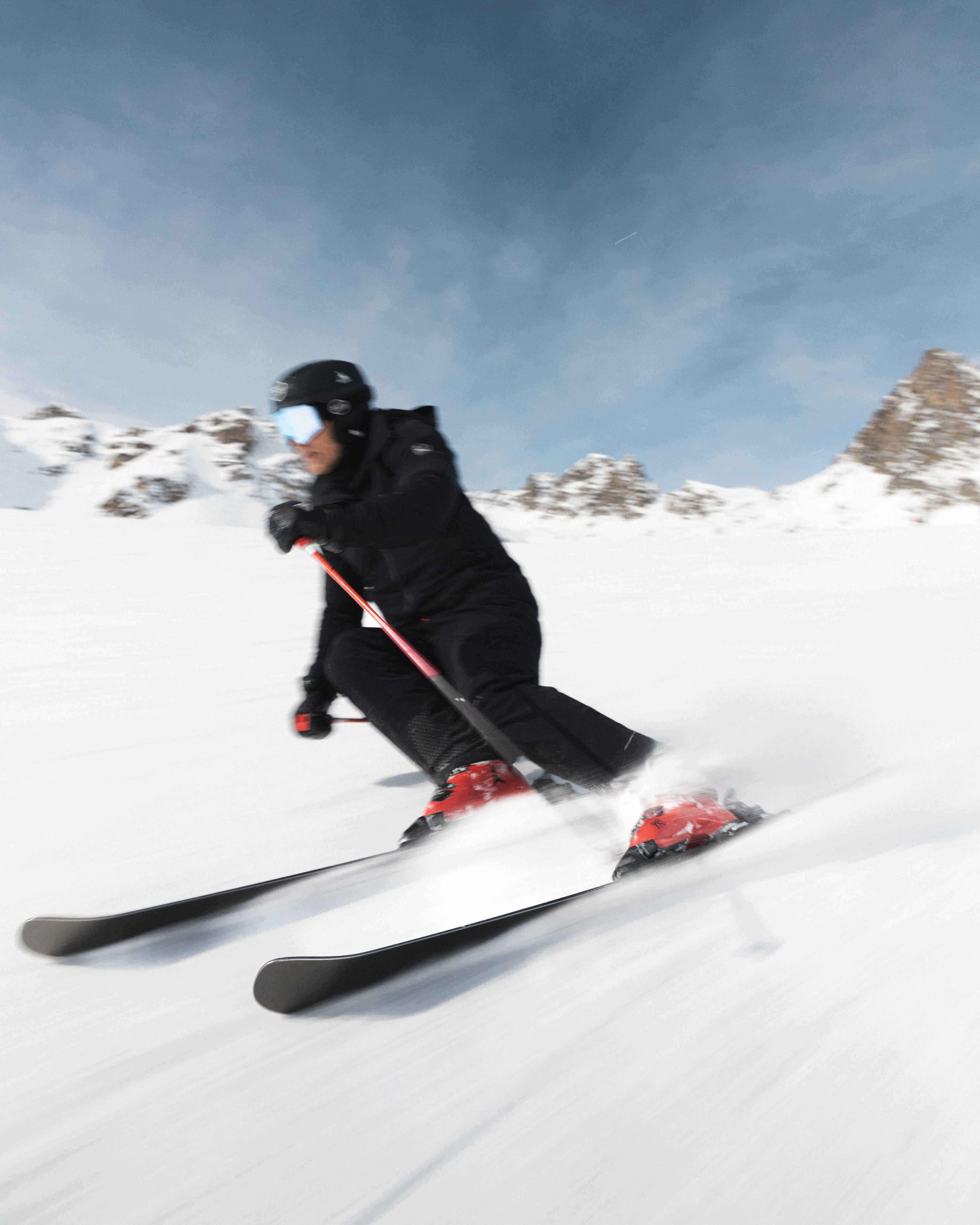 A skier in a black outfit with red ski boots carving through deep snow under a blue sky, with rocky mountains in the background.