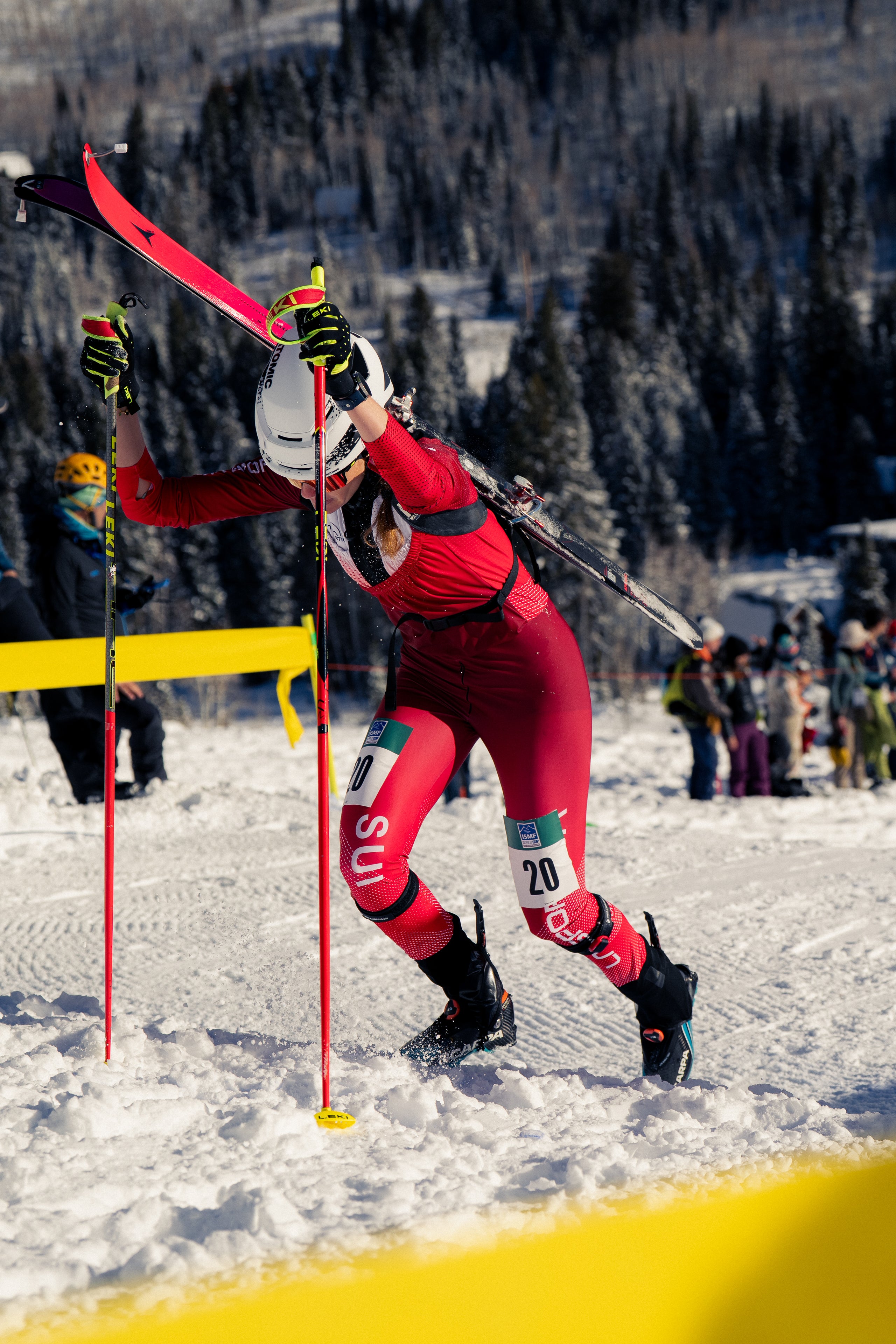Alessandra Schmidt in a red and black ski suit, wearing a white helmet and holding ski poles and skis, captured mid-action on a snowy mountain slope with a forest background.
