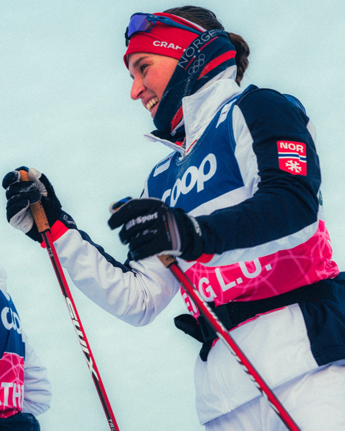 Two women cross-country skiing outdoors in bright snow, wearing matching navy, white, and pink ski uniforms with visible sponsor logos and a Norwegian flag patch.
