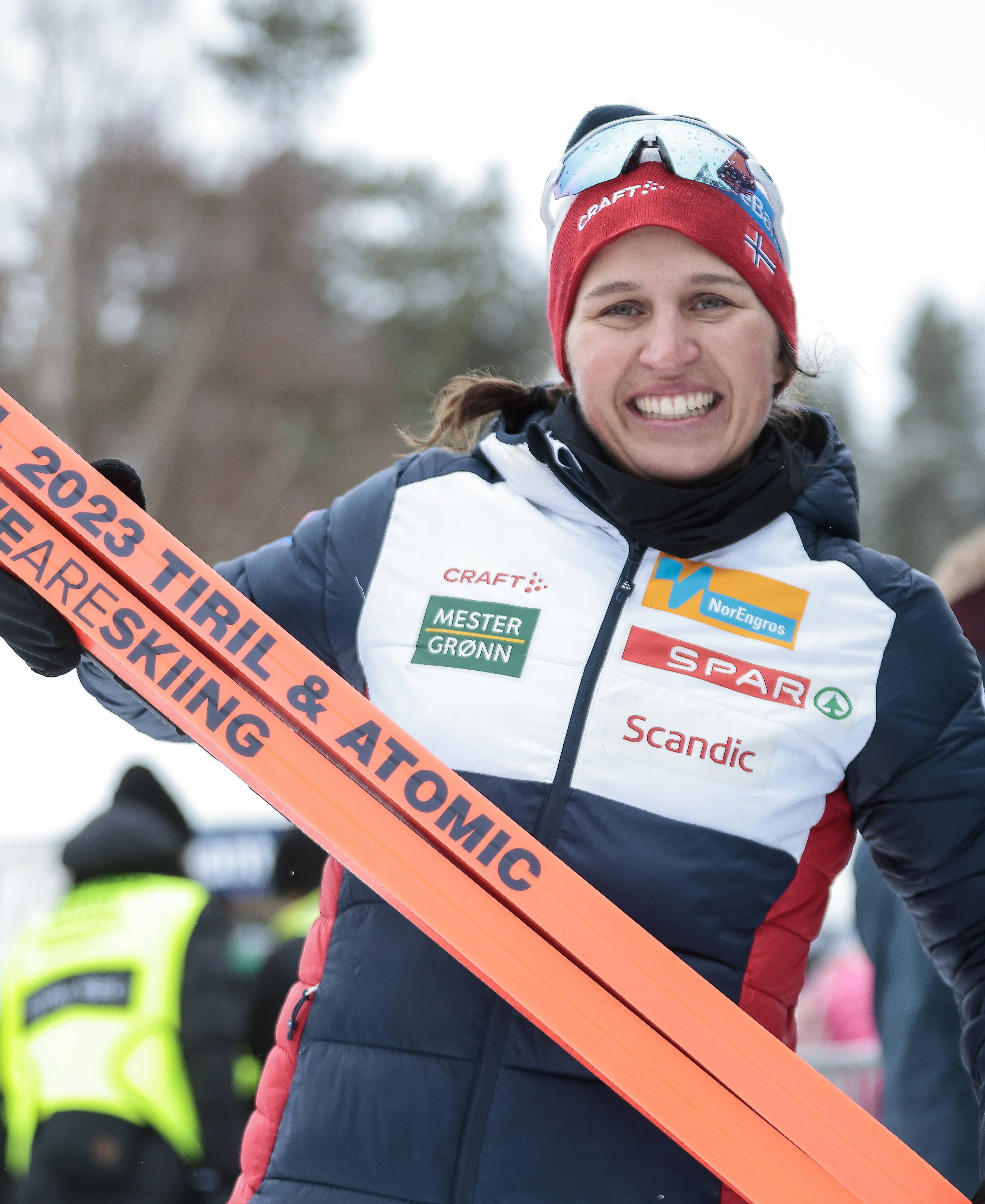 A smiling Tiril Udnes Weng in a white, navy, and red ski jumping jacket holds an orange ski across the frame. The jacket features sponsor logos.