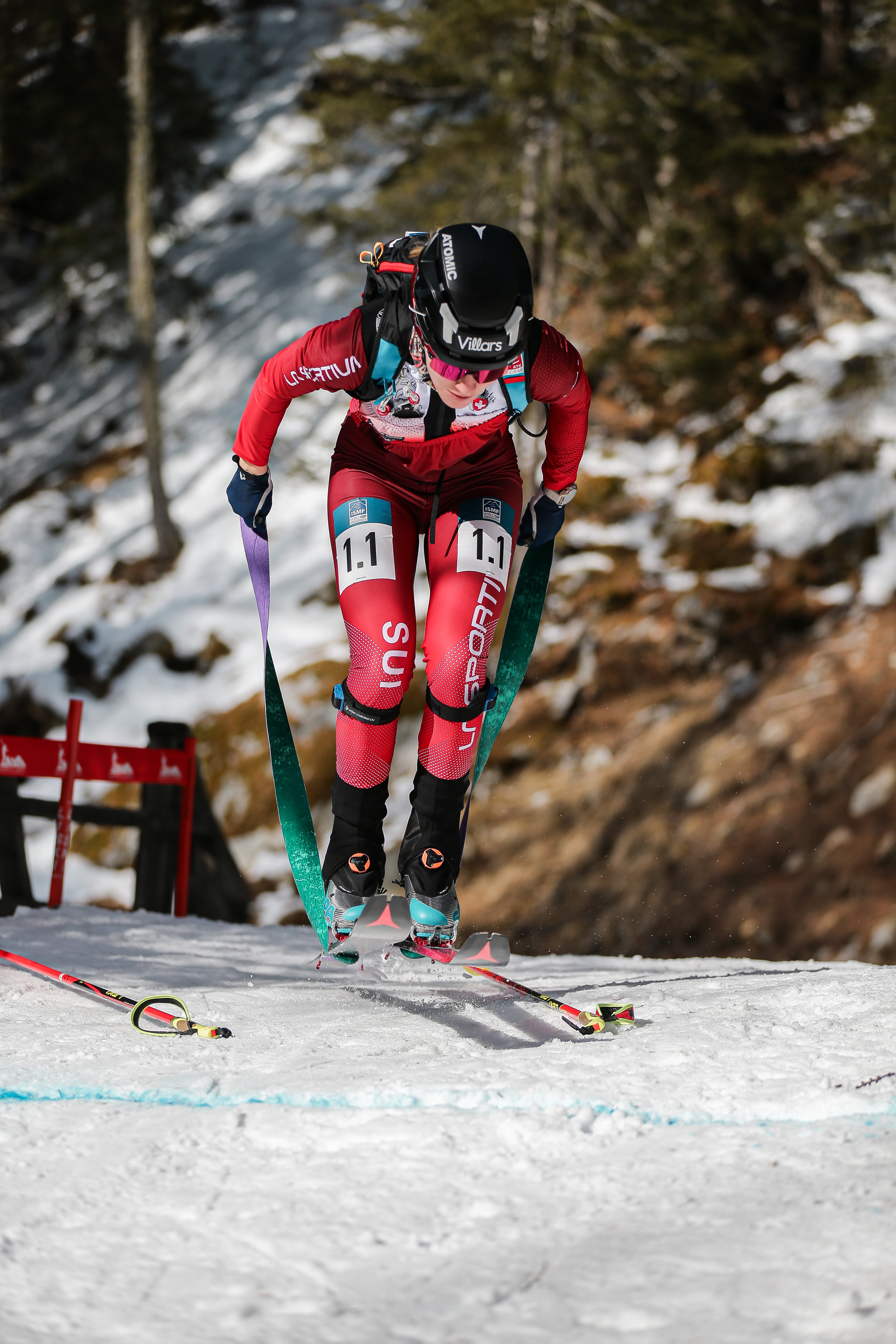 Caroline Ulrich in a red and pink suit  during a skin transition on a snowy mountain course, with trees and rocks in the background.