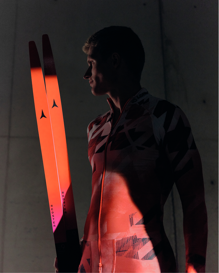 Mika Vermeulen profile wearing a dark patterned ski suit and holding bright orange ski tips against a dim concrete wall.