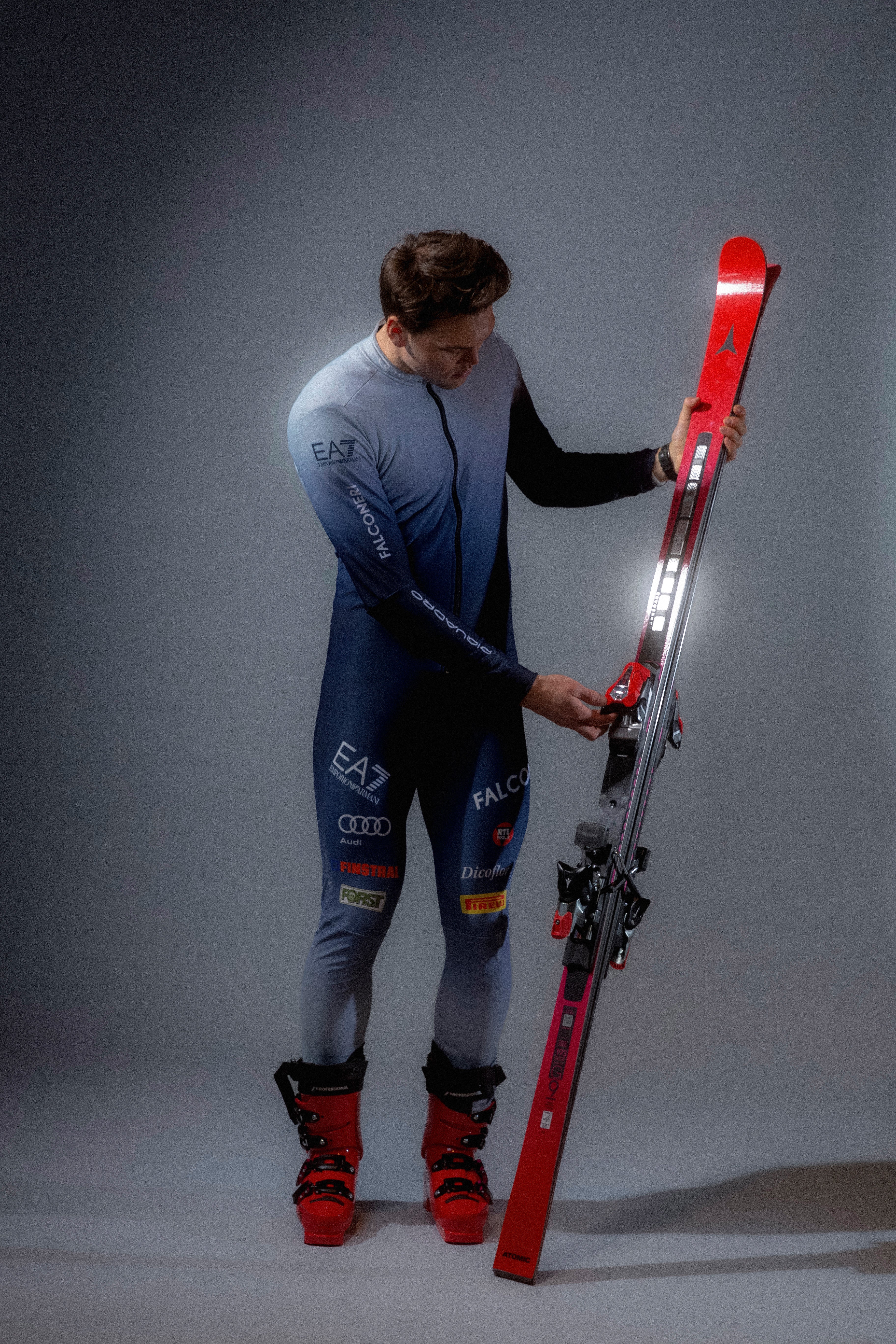 Alex VInatzer in a blue and gray ski suit adjusts the bindings on a red and silver ski, standing against a dark gray studio background.