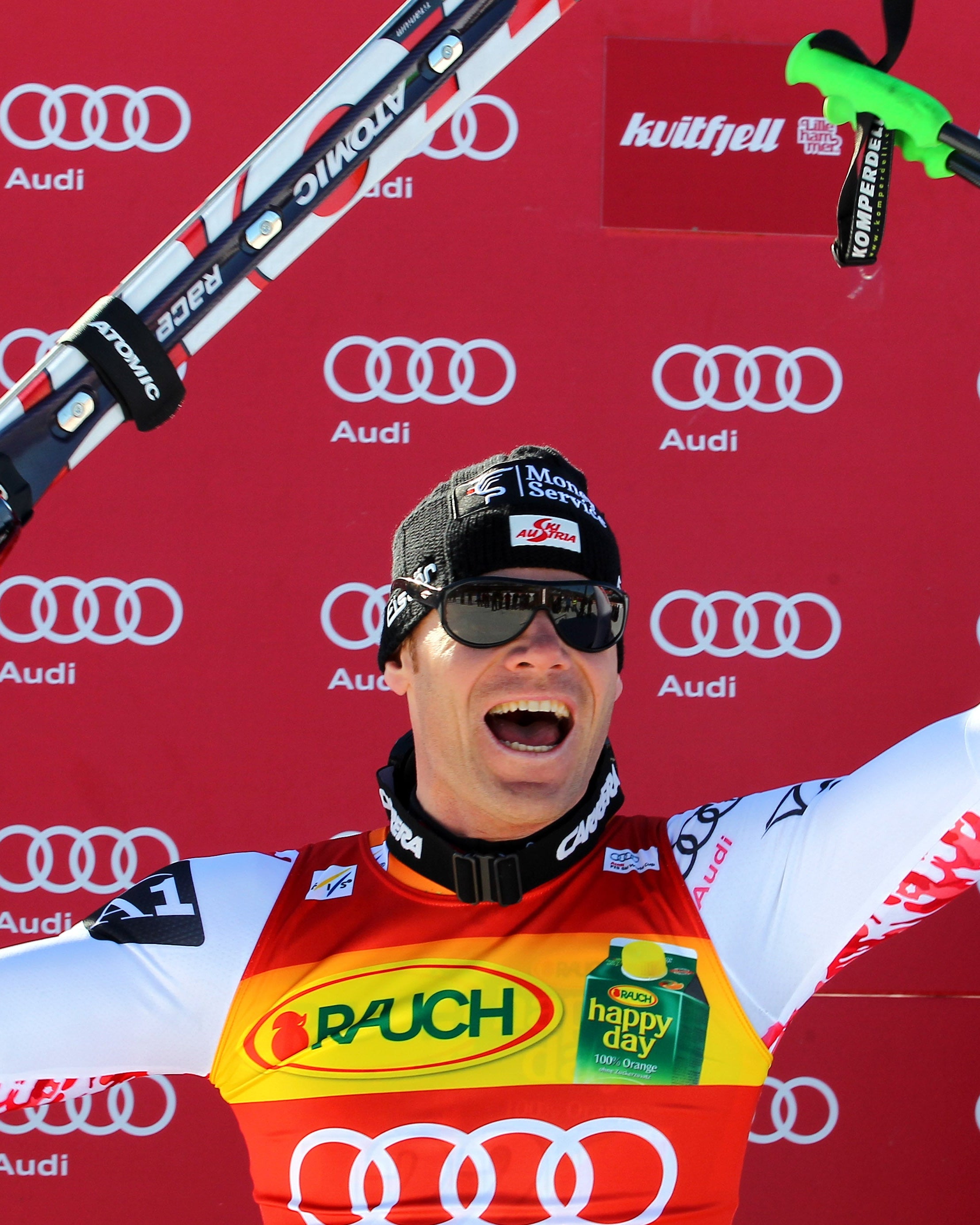 Michael Walchhofer in a red and white racing suit celebrates on a podium, holding ski poles and skis against a red  backdrop.