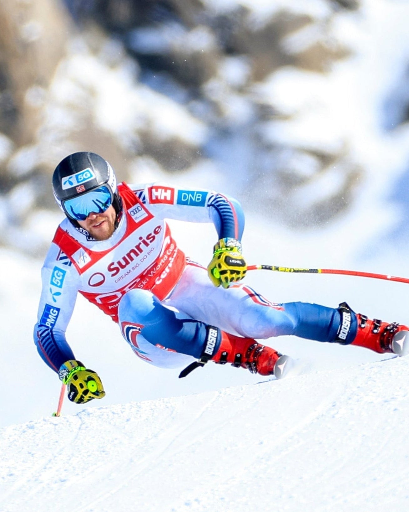 Aleksander Aamodt Kilde in a red, white, and blue racing suit carving through fresh powder on a snowy mountain slope, with rocky peaks blurred in the background.