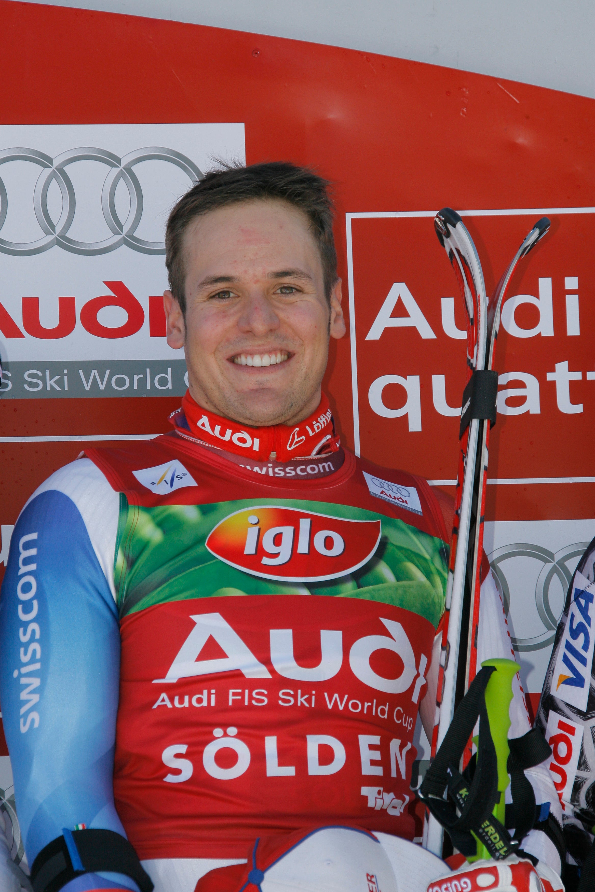 A smiley Daniel Albrecht in a red, green, and blue racing suit, holding ski boots and skis in front of an Audi sponsor backdrop.
