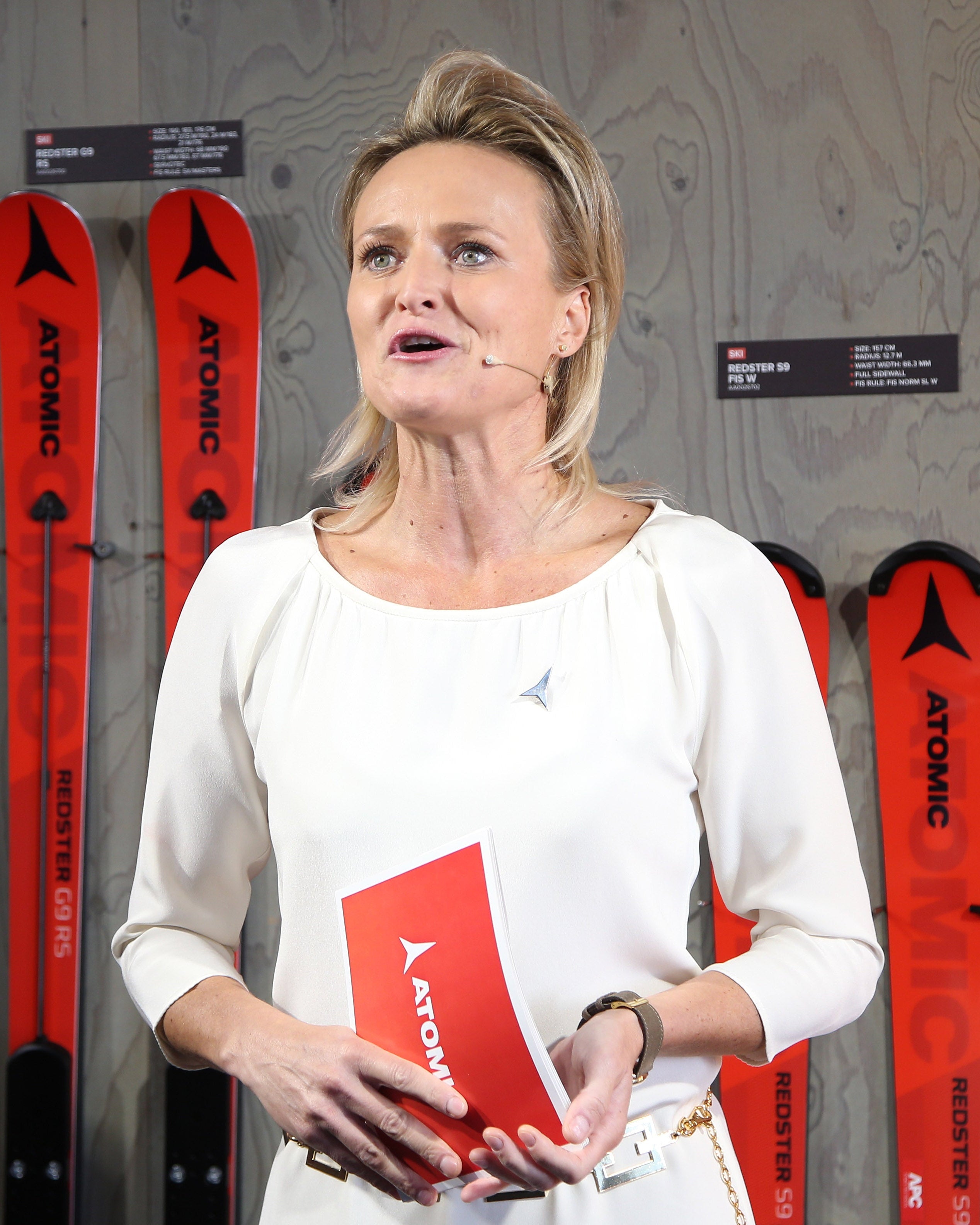 Alexandra Meissnitzer in a white blouse stands in front of a wood-paneled wall holding a red booklet, with several red Atomic skis mounted behind her.