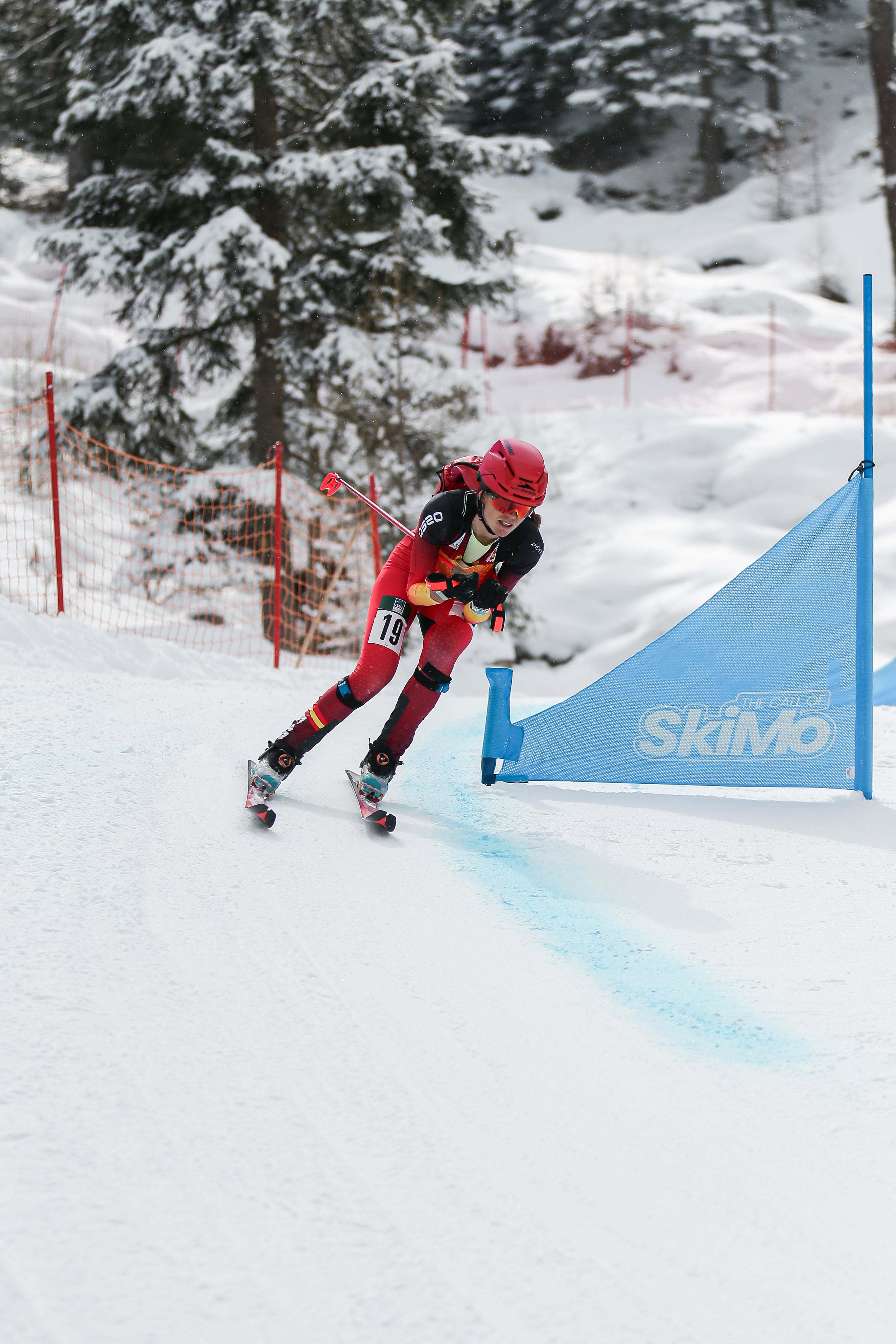 Ana Alonso in a red and black outfit with a red helmet navigates a snowy slope past a blue SkiMo gate, with snow-covered trees in the background.