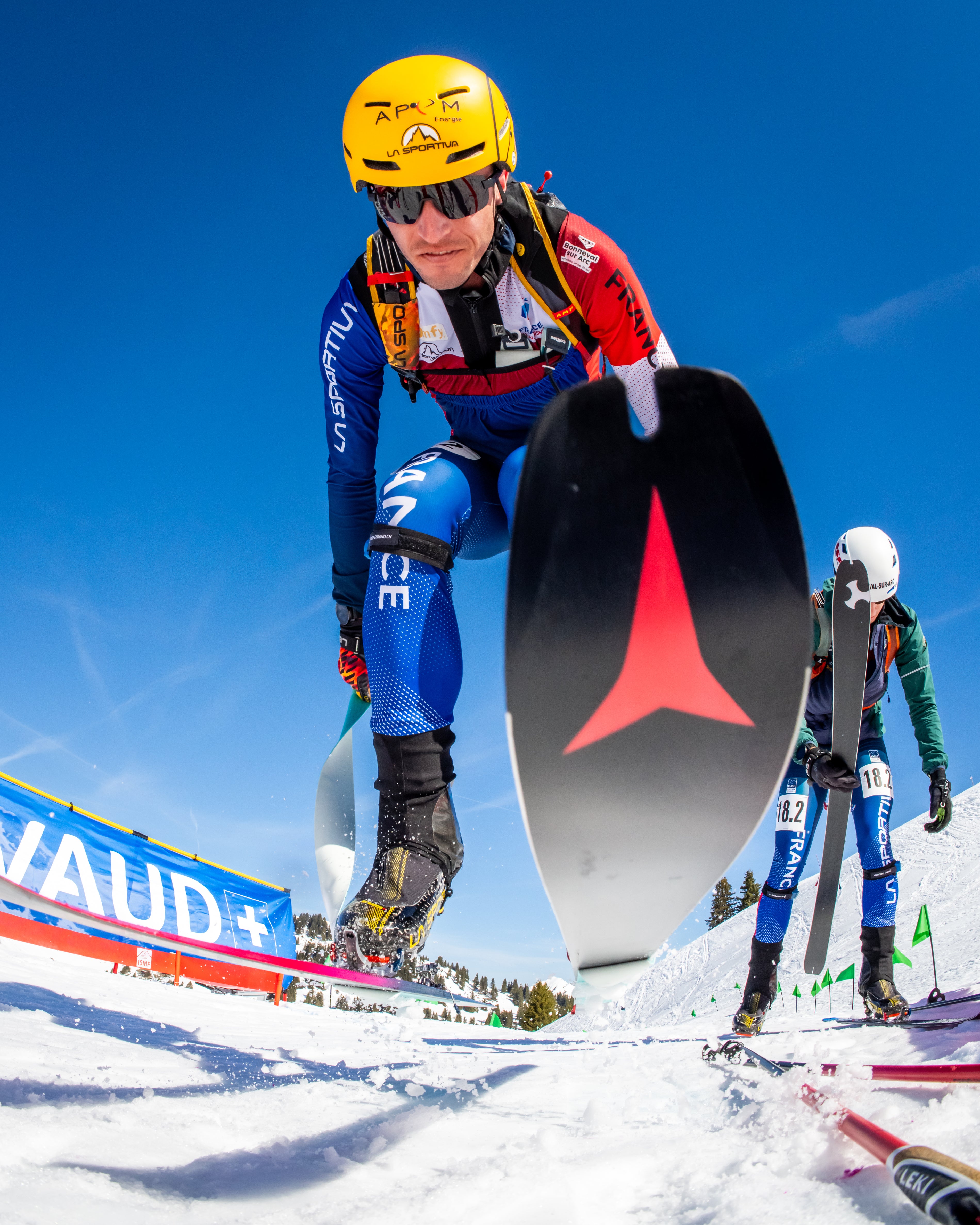 Thibault Aselmet in a blue, red, and white racing suit with a yellow helmet and sunglasses during a race. 