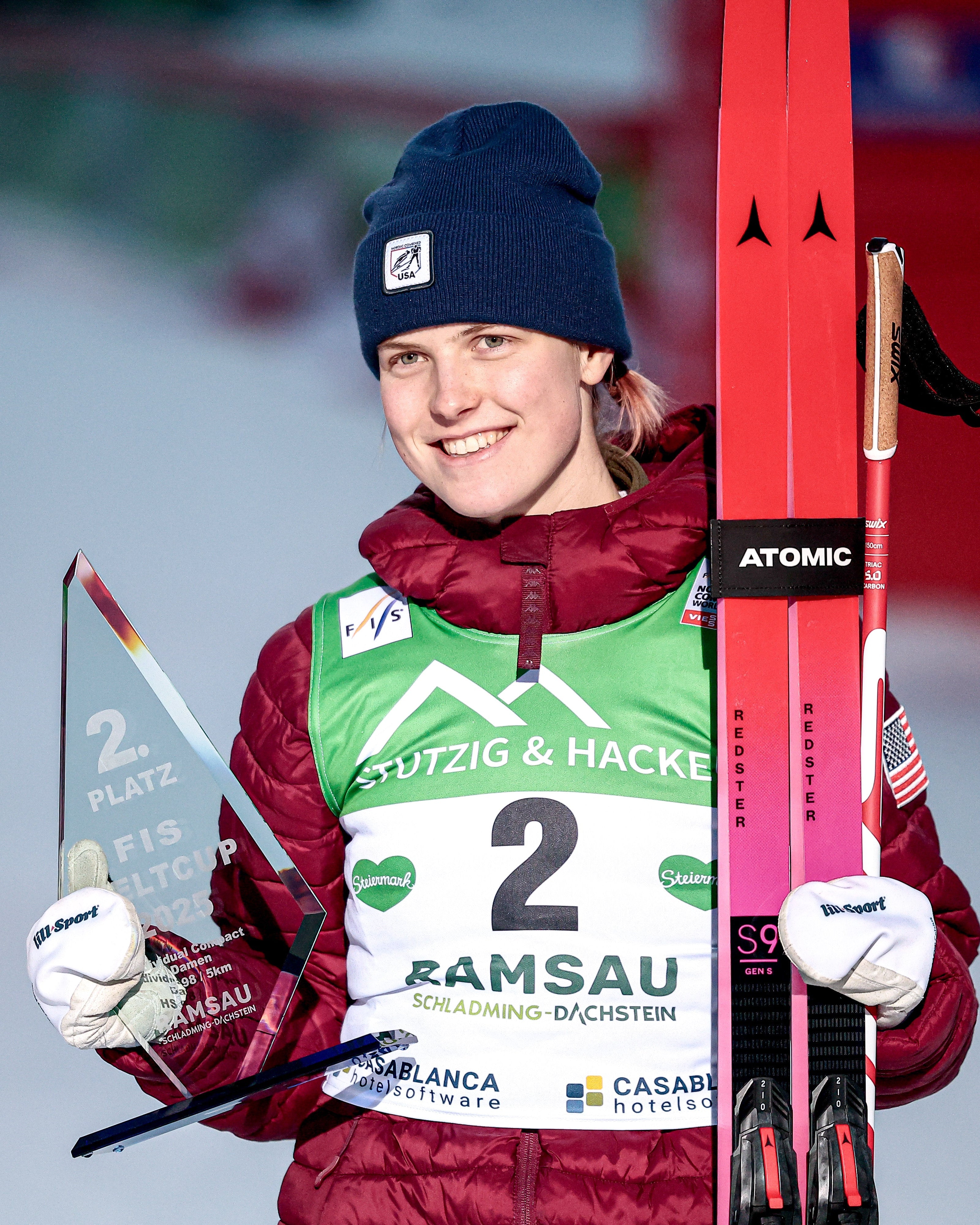 The nordic ski racer Alexa Brabec wearing a dark blue beanie, a red and green jacket, and white gloves, holding a glass award trophy and red skis with 'ATOMIC' branding on a snowy podium.