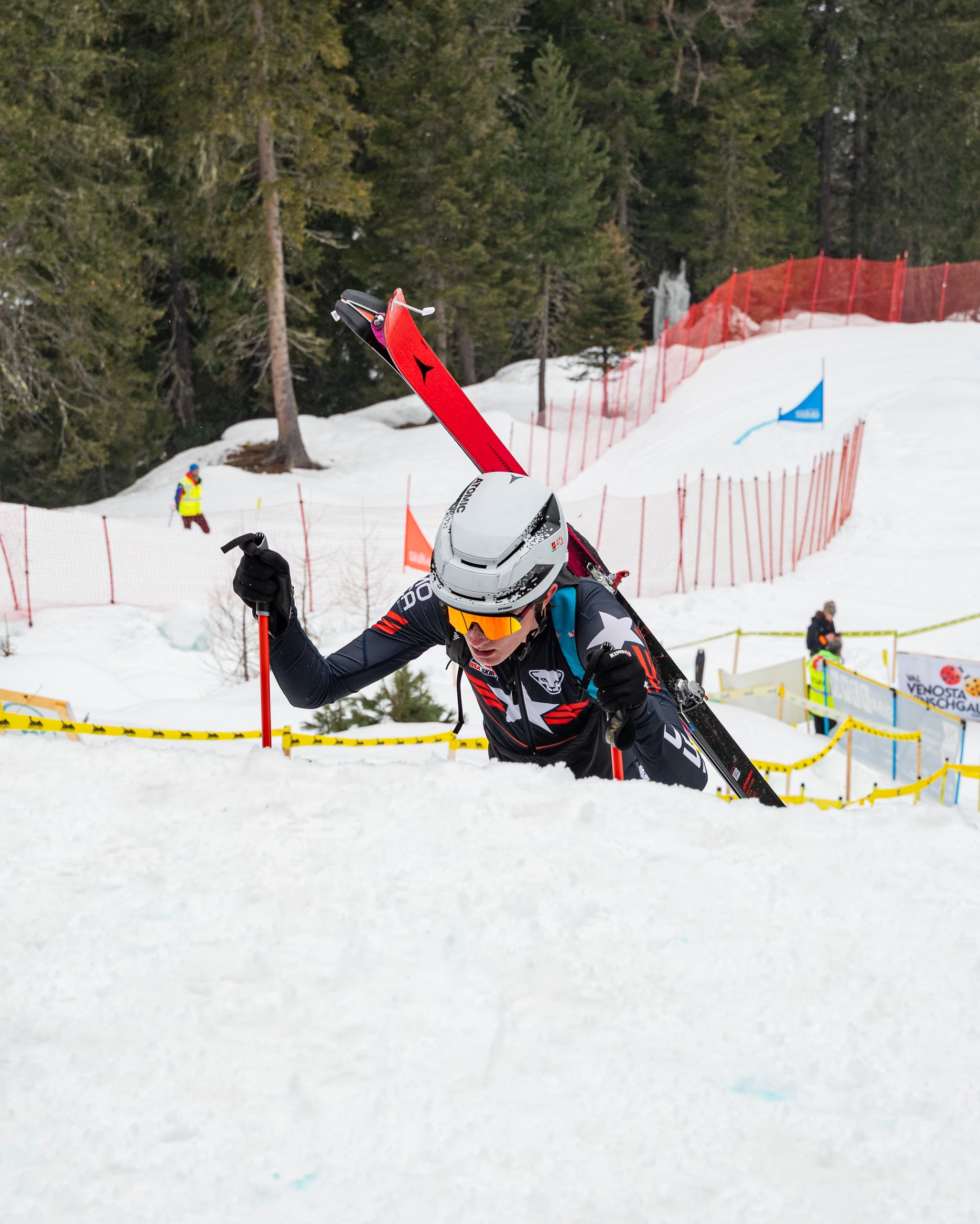 Griffin Briley in a black and white racing suit with a white helmet and goggles, holding ski poles and skis, climbing a snowy slope with orange safety netting and pine trees in the background.