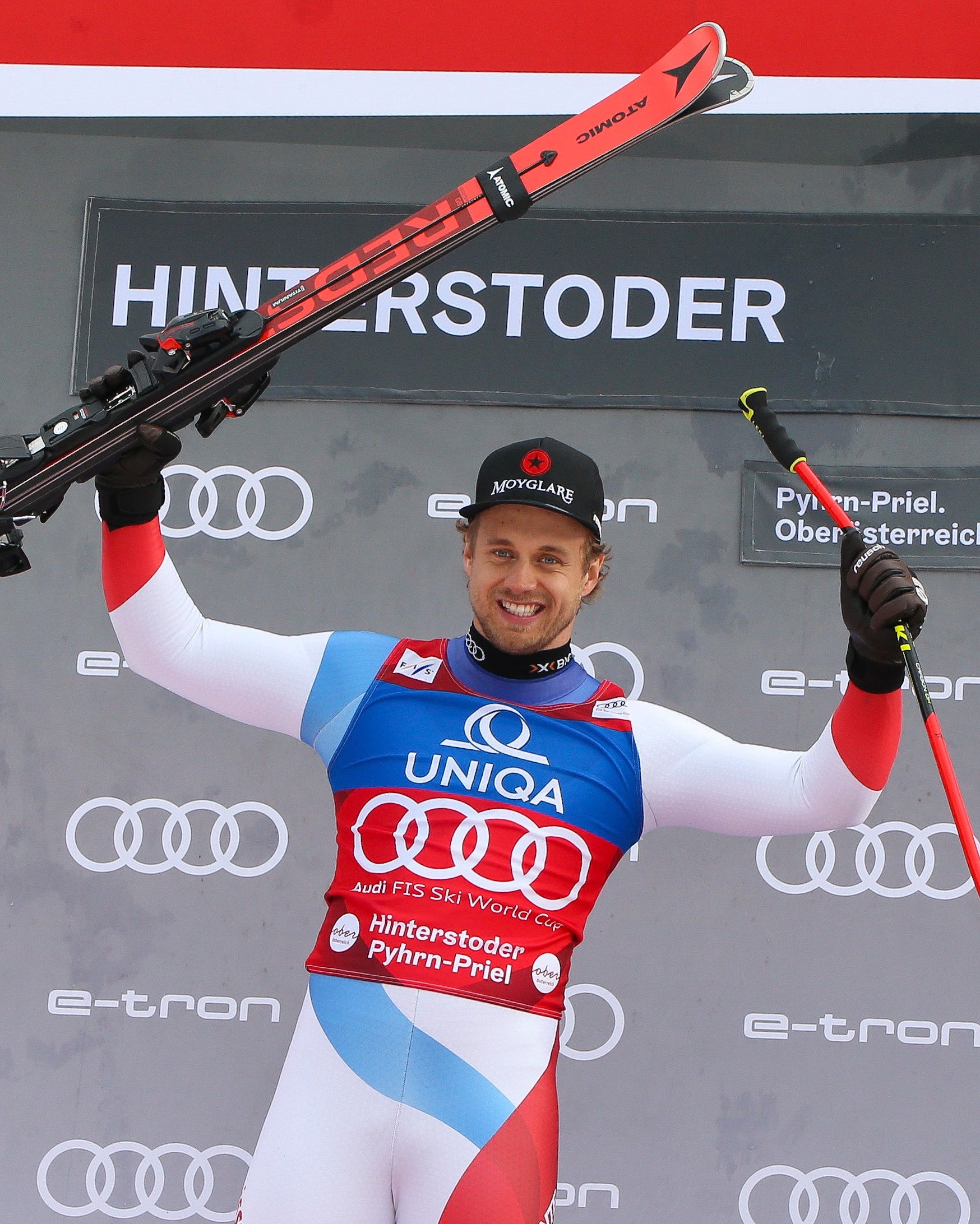 Mauro Caviezel in a red, white, and blue racing suit stands on a podium holding red skis and ski poles in front of a grey backdrop.