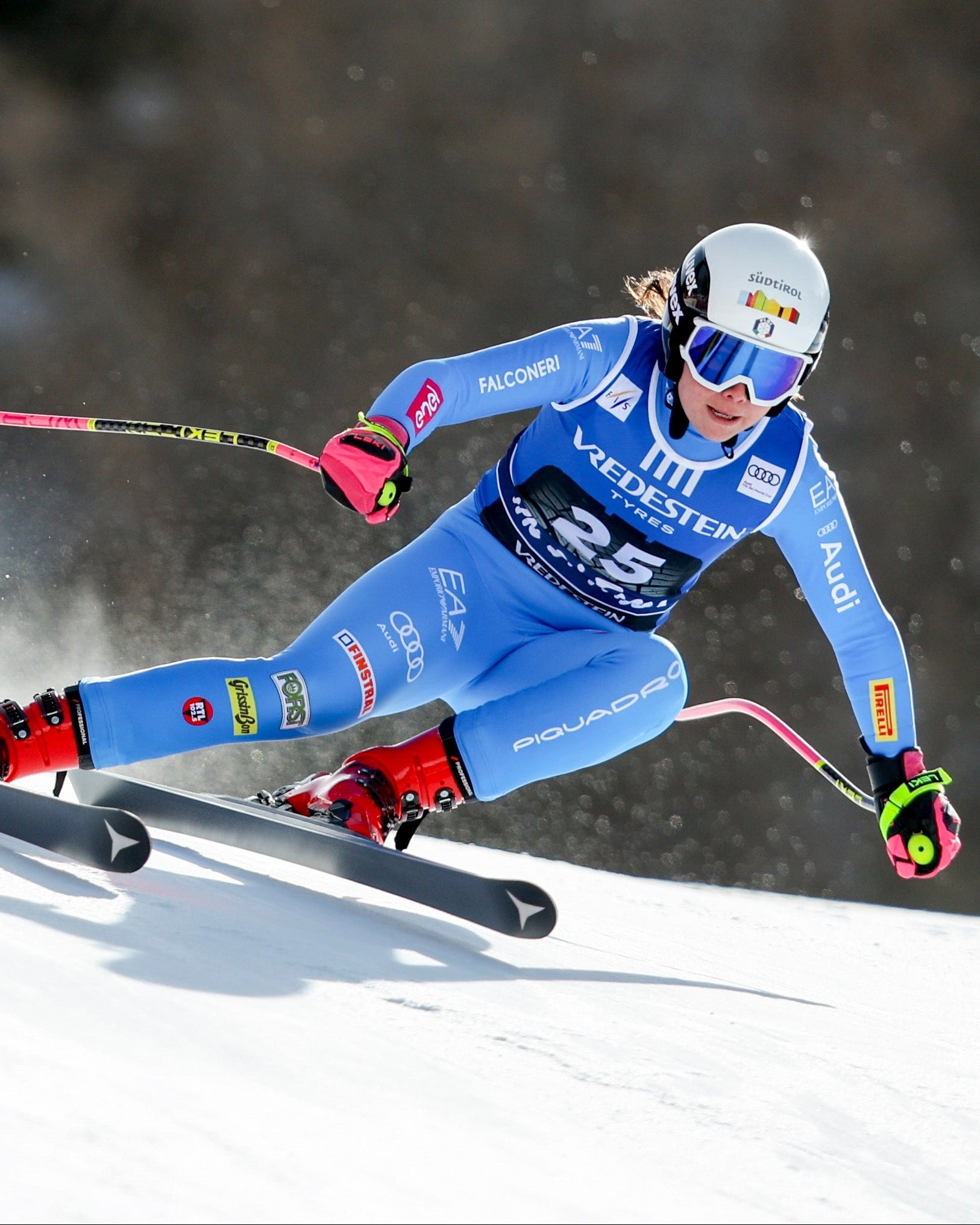 Nadia Delago in a blue and white racing suit with red boots leans sharply into a turn on a snowy race slope, kicking up snow spray, with dark trees blurred in the background.
