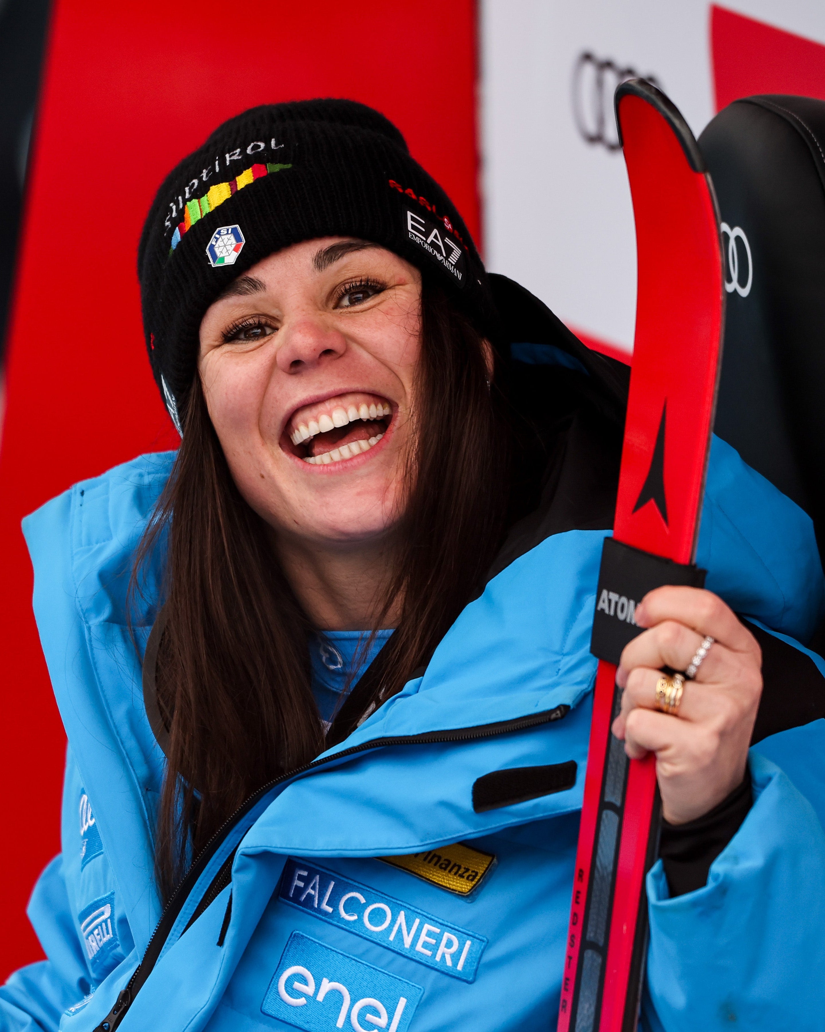 Nicol Delago smiling in ski resort attire wearing a bright blue jacket and black beanie, holding red skis with sponsor logos in the background.