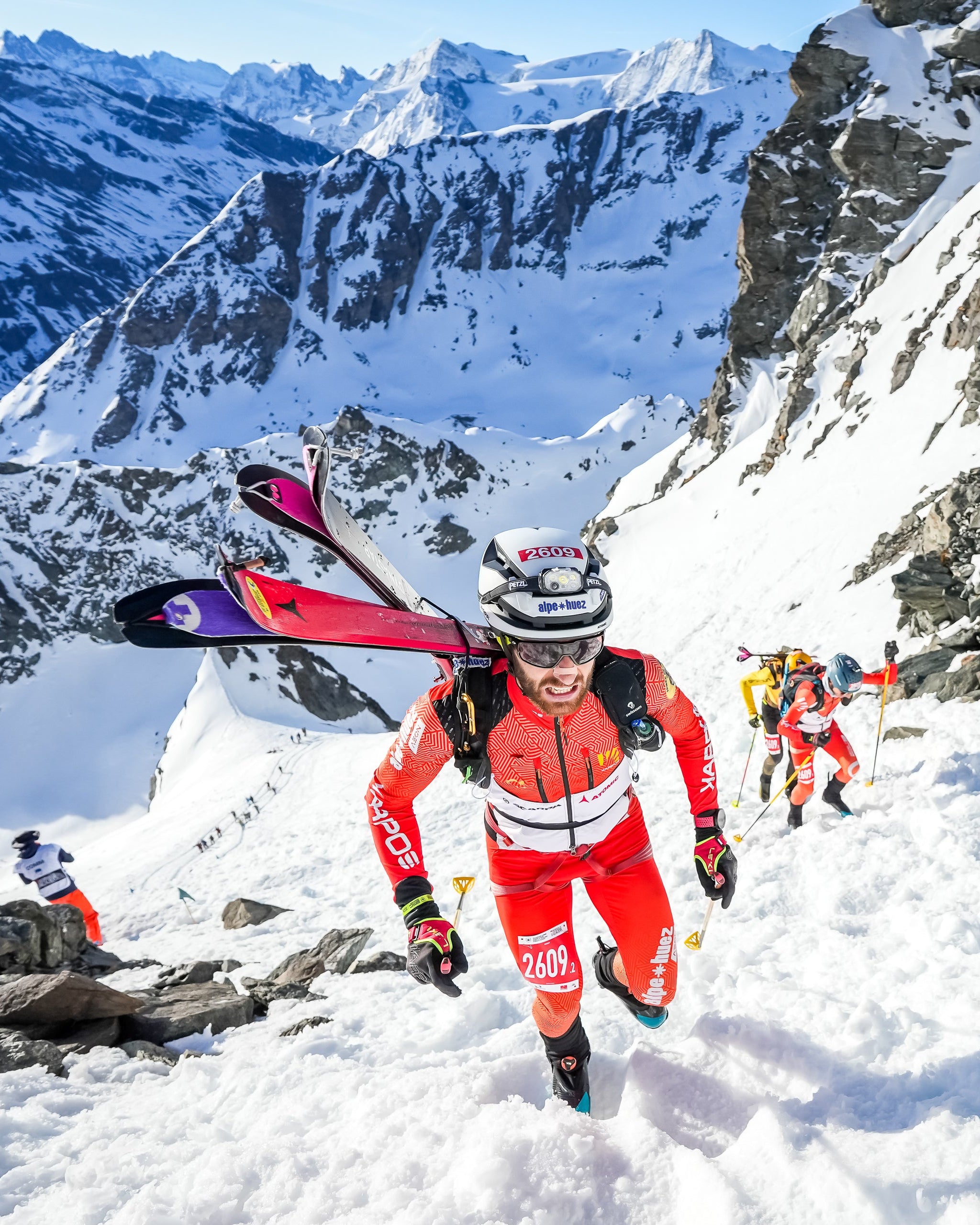 Samuel Equy in a bright red and white ski suit carries skis over his shoulder while walking through deep snow on a steep mountain slope, with snow-covered peaks and rocky cliffs in the background.