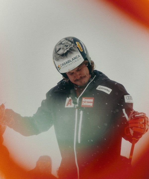 Manuel Feller in a snowy outdoor setting wearing a black ski jacket with sponsor logos and a patterned helmet.