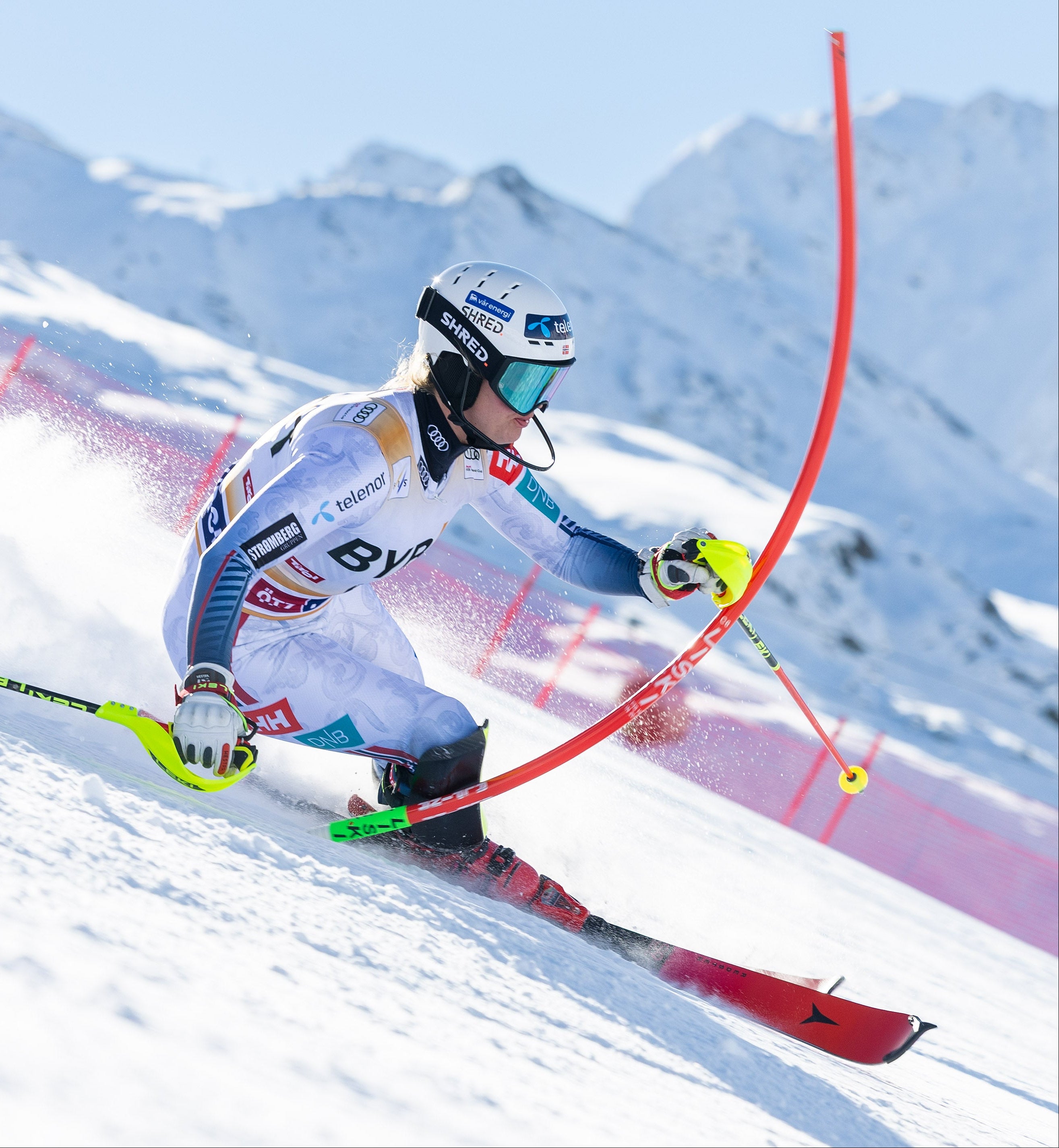 Mina Holtmann a white and blue racing suit navigates a slalom course during an alpine race, with snow-covered peaks in the background.