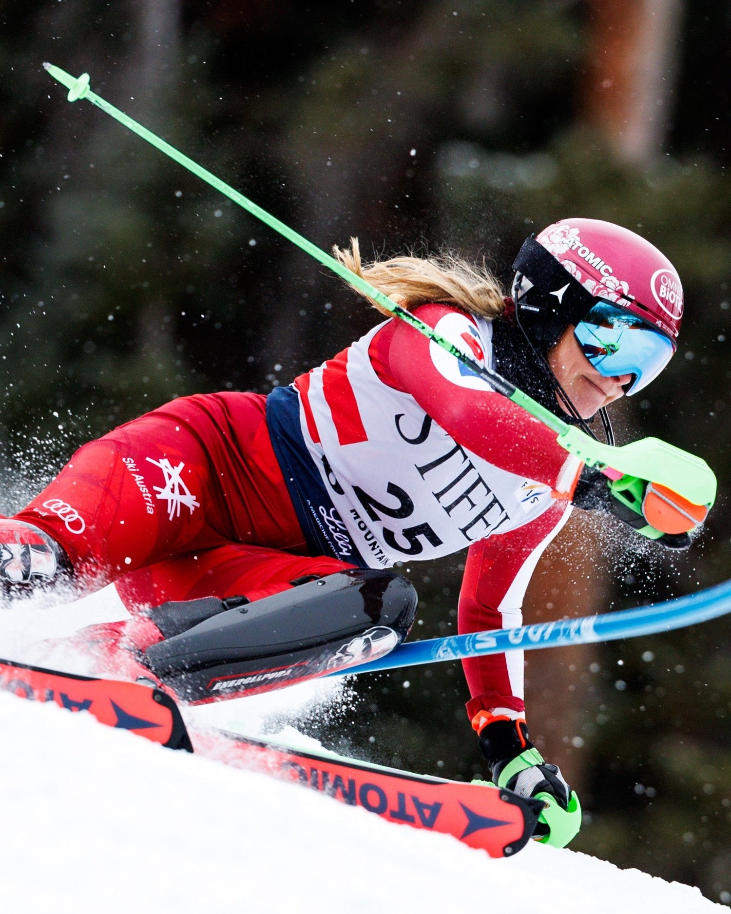Katharina Gallhuber in a red and white racing suit leans sharply into a turn on snow, with ski spray kicking up behind her. The skier wears a red helmet, blue goggles, and holds green and blue poles.