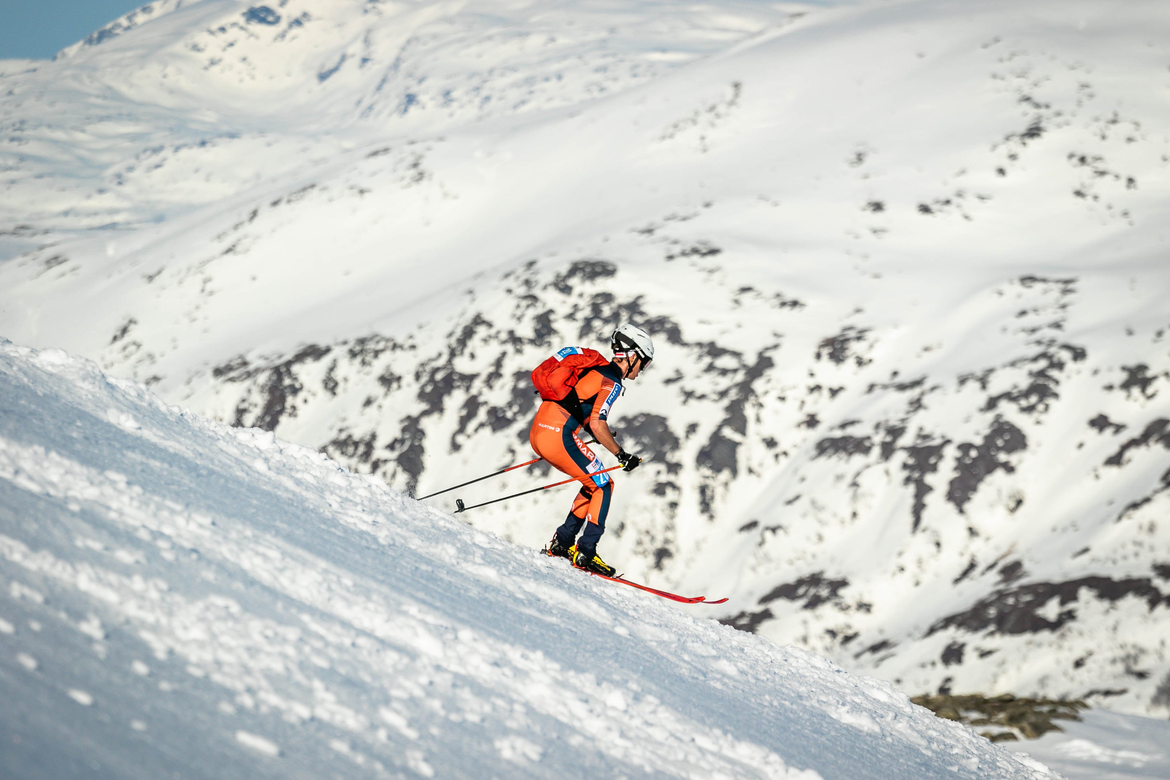 Daniel Ganahl in an orange and black suit navigates a snowy mountain slope with snow-covered peaks in the background.