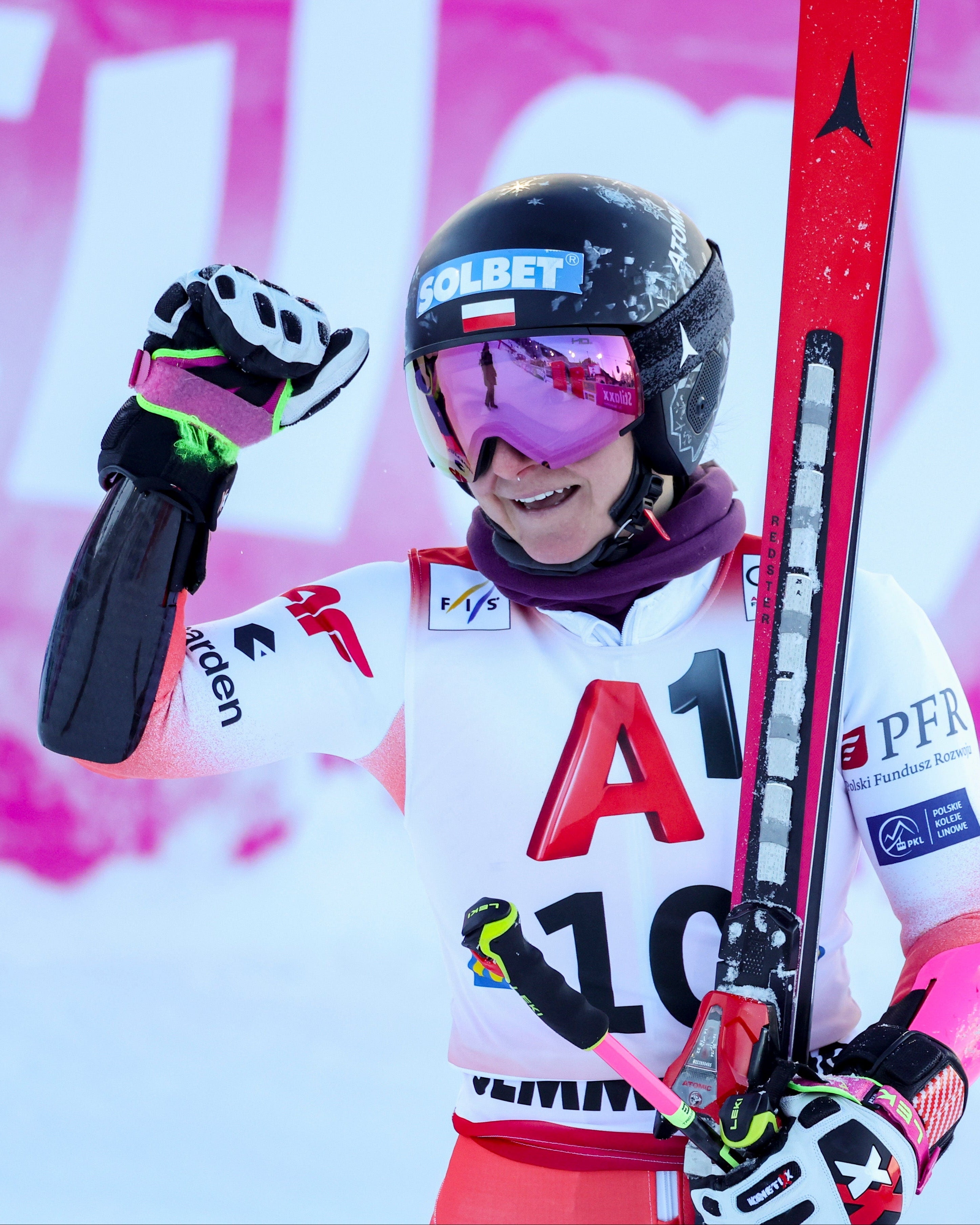 Maryna Gasienica-Daniel celebrates on the snow after a run, wearing a white and red competition suit with the number 10, a black helmet, and holding red skis. The background features a pink and white banner.