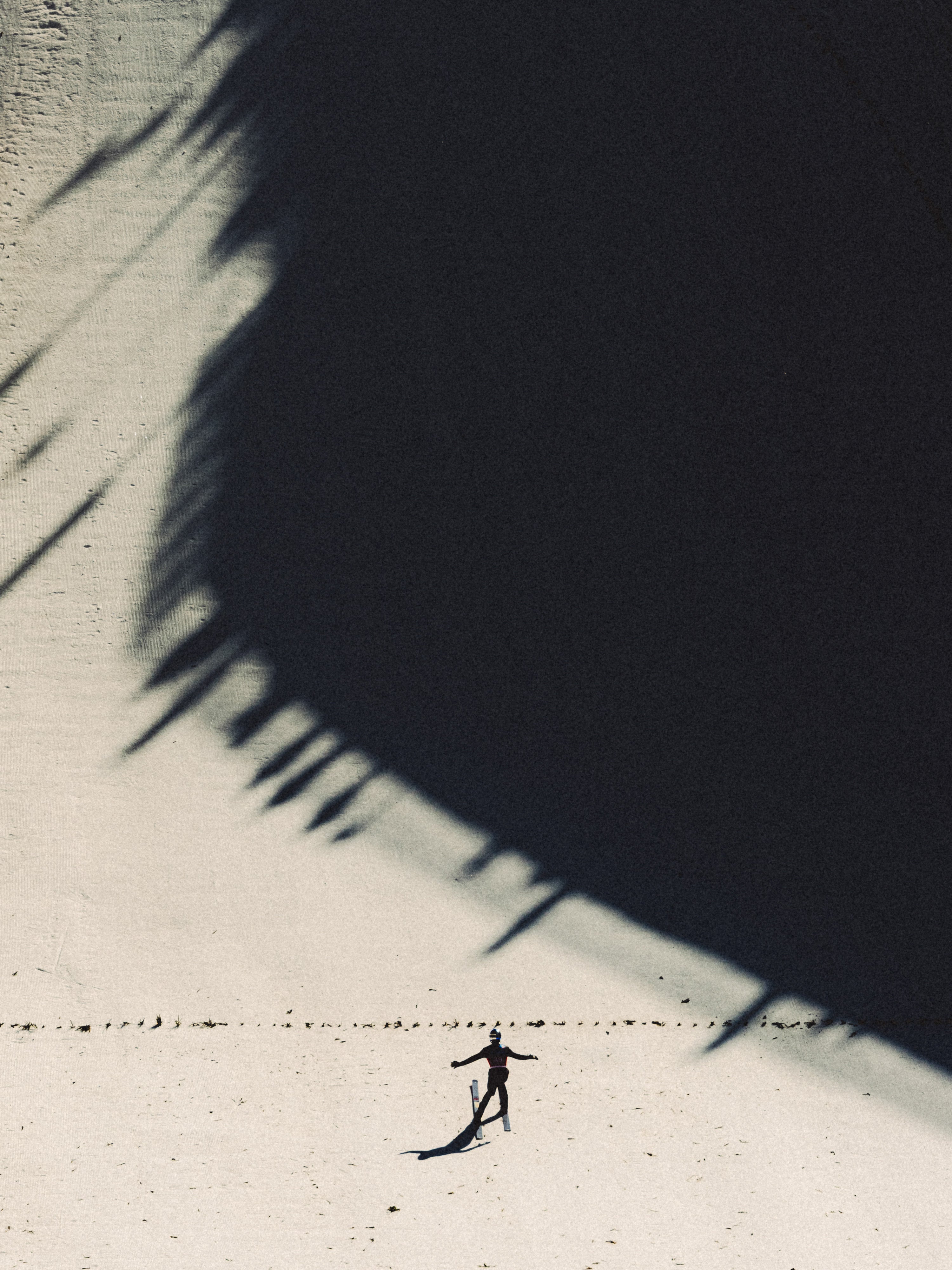 A minimalist photograph of a tiny Vinzenz Geiger standing on a light snowy ground, casting a long shadow that stretches across a dark, shadow-filled area under bright sunlight.