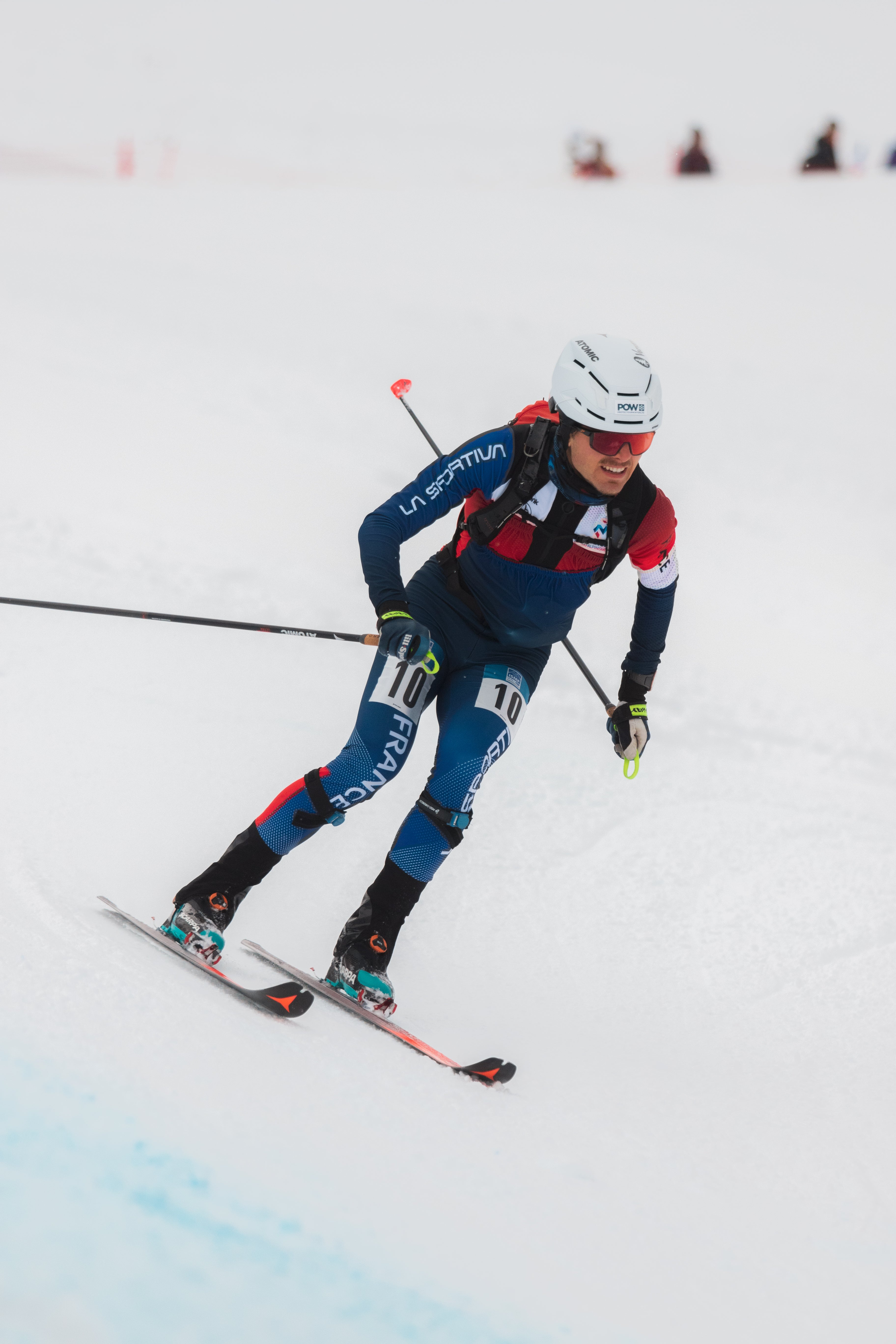 Pablo Giner in a blue, red, and white racing suit leans into a turn on a snowy slope, holding ski poles and wearing a white helmet and sunglasses.