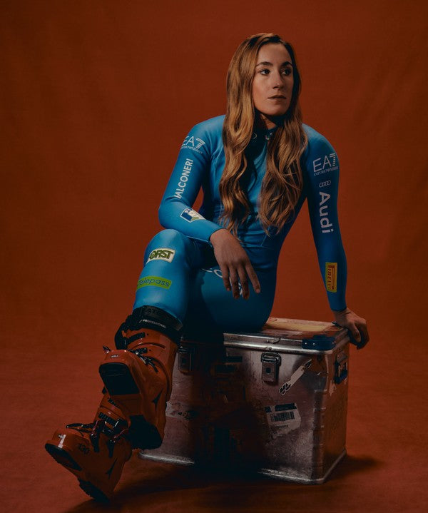 Soffia Goggia in a blue racing suits sits on a metal trunk against a warm orange-brown studio backdrop. She is wearing brown racing boots, and the suit features visible sponsor logos.