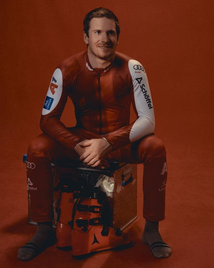 Fabio Gstrein wearing a red racing suit with white sponsor sleeves sits on a box against a solid red-brown studio background. Visible logos on the left sleeve.