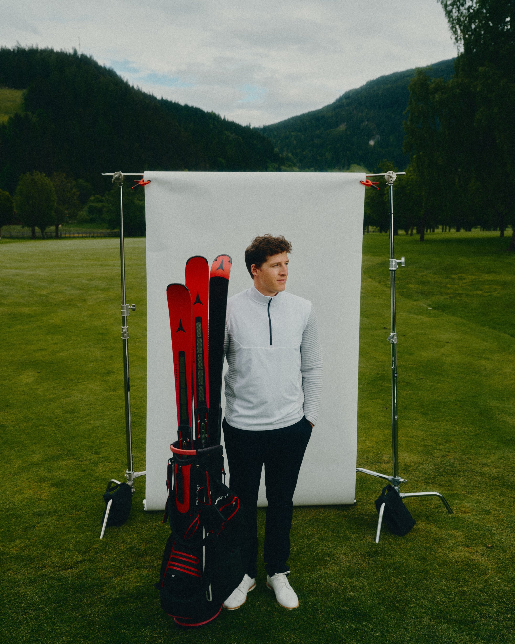Raphael Haaser stands on a grassy golf field in front of a white studio backdrop stand, wearing a light gray quarter-zip pullover, dark pants, and white shoes. A black and red golf bag with red skis in it leans against him, with mountains in the background.