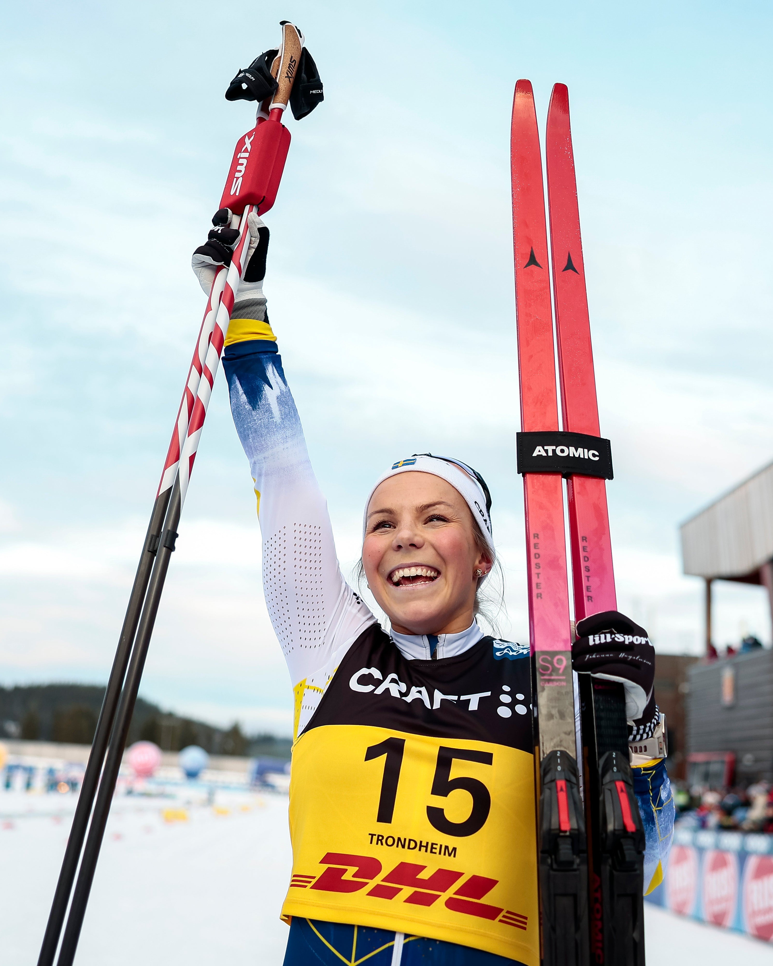 Johanna Hagstroem celebrates in the snow, holding up her ski pole with a black and red flag while holding a pair of red skis. She is wearing a racing suit with a yellow bib numbered 15.