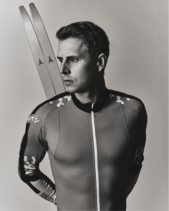 A black and white studio portrait of Simen Hegstad Krüger wearing a fitted racing suit and holding a pair of skis, with a light gray background.