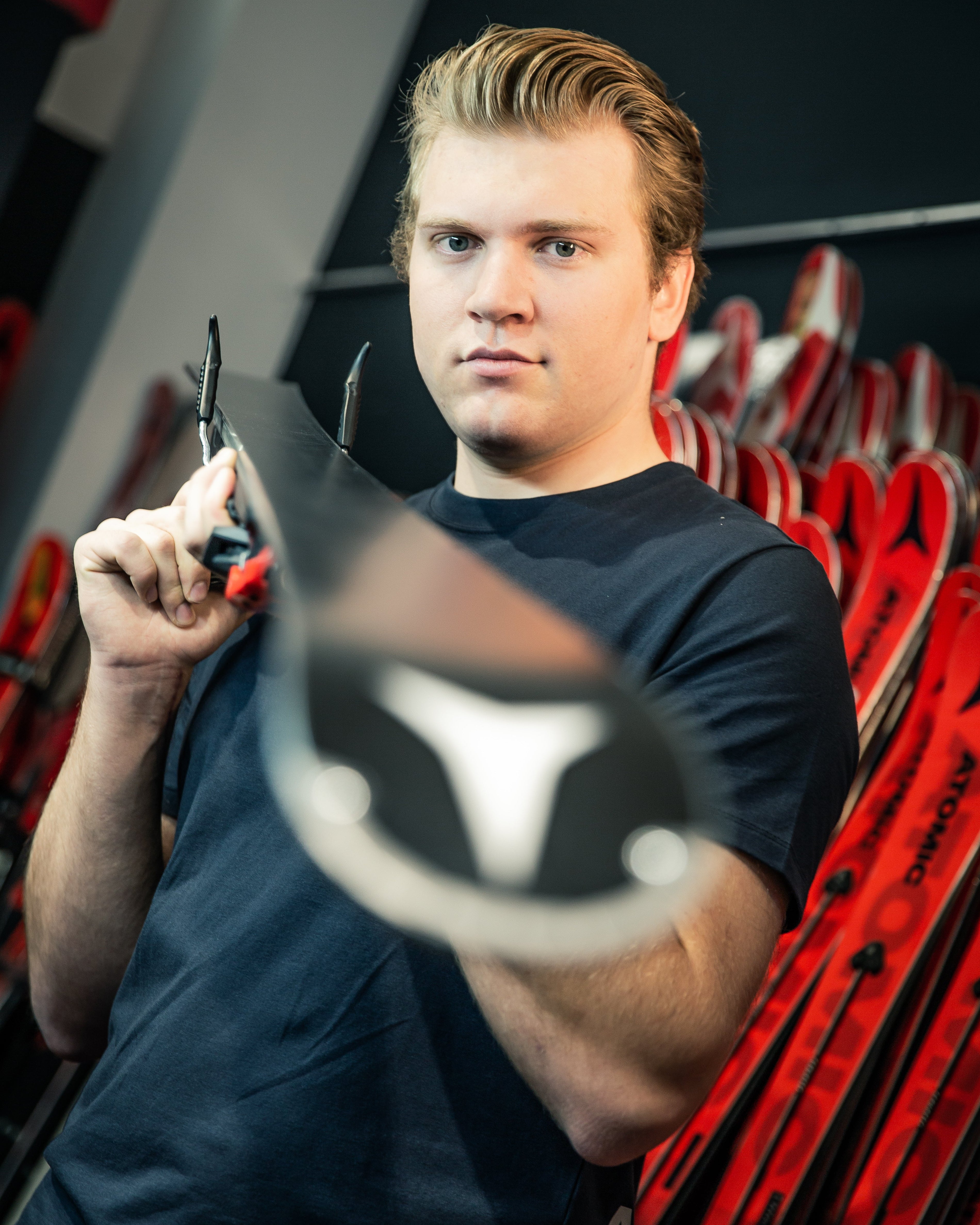 Niels Hintermann in a dark navy t-shirt holds a ski boot in the foreground with pliers, with a rack of red and black skis in the background.